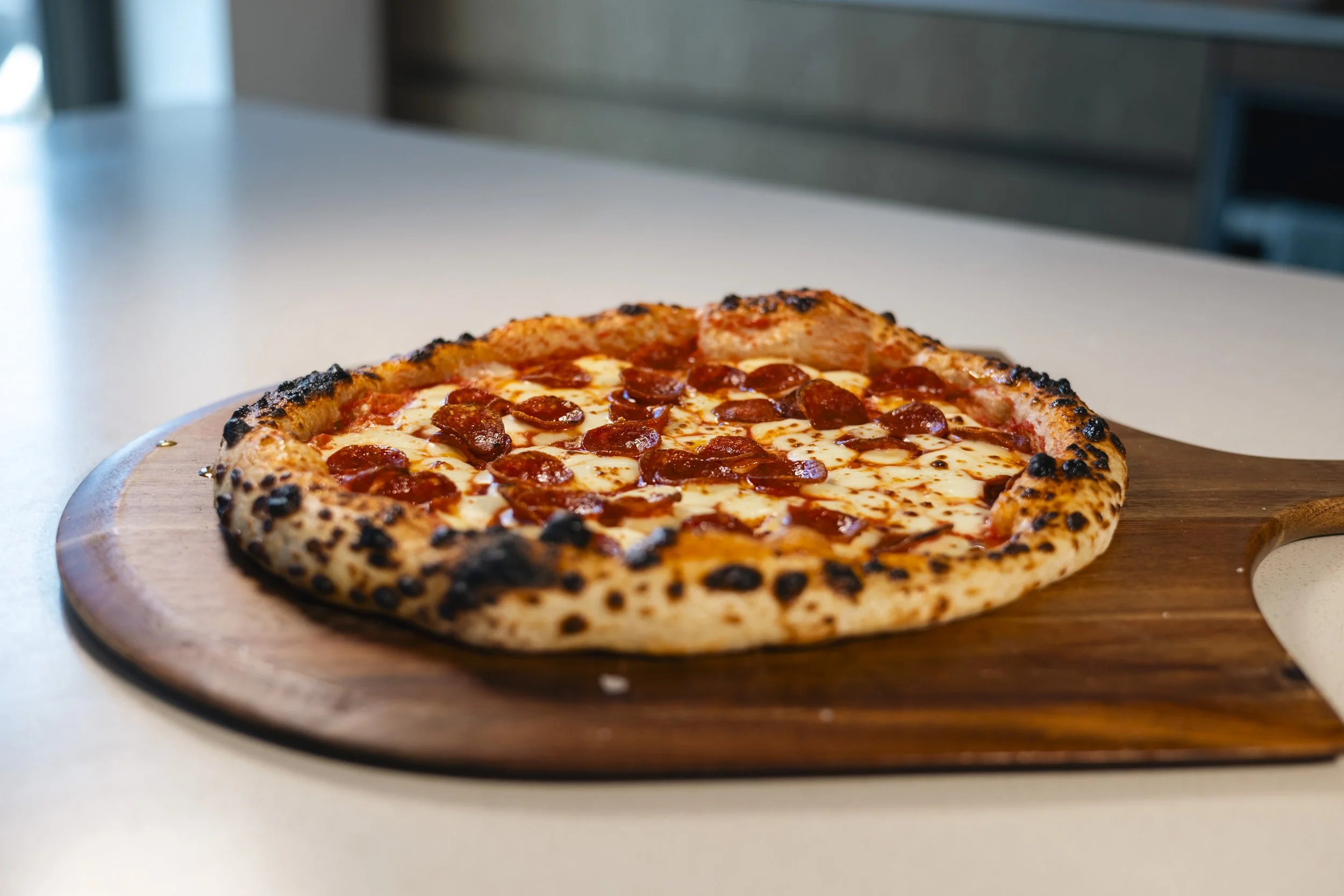 A pepperoni pizza with a charred crust resting on a wooden pizza peel on a white countertop.