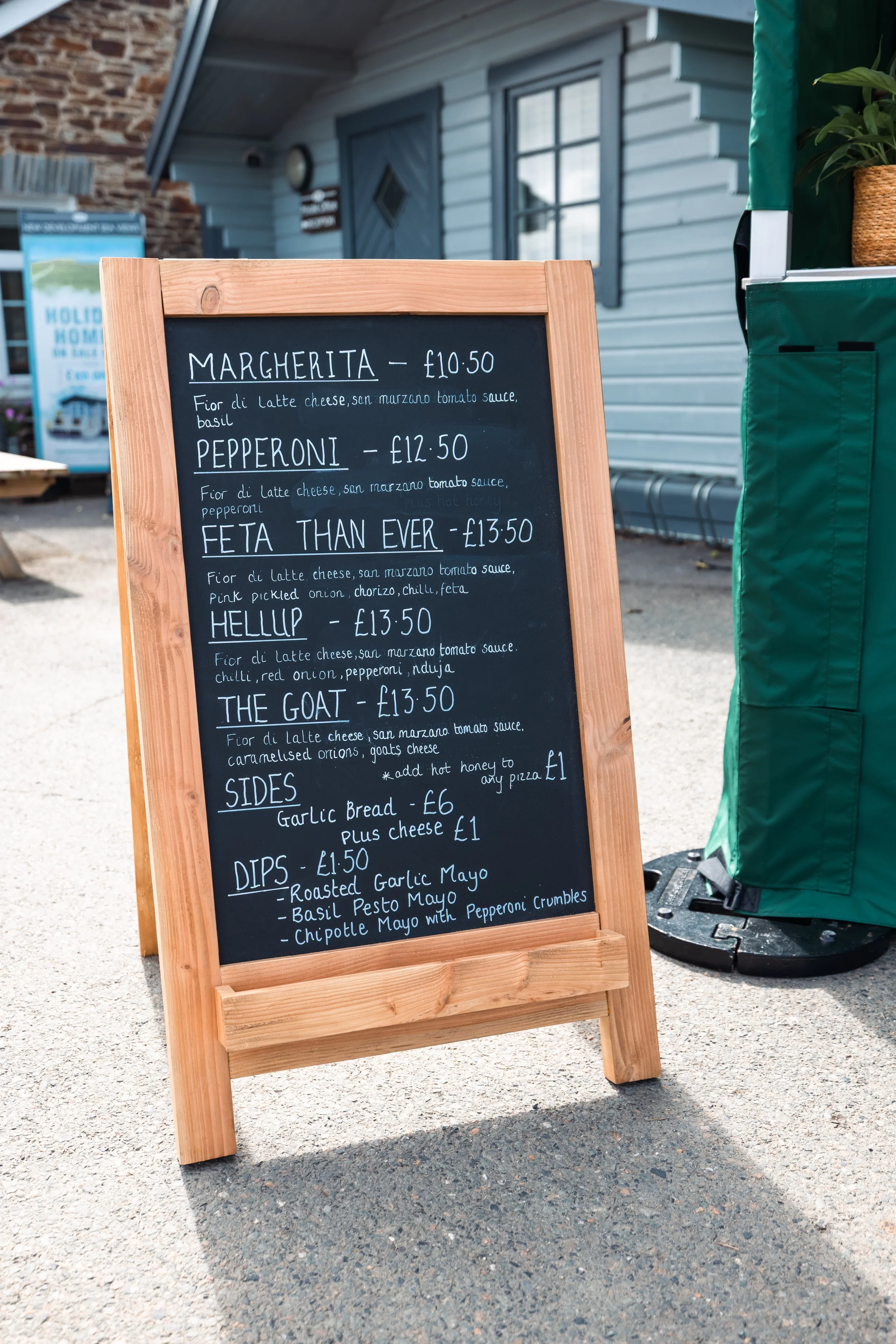 A blackboard menu on a wooden stand outside a building, listing various pizza options and prices in American English.