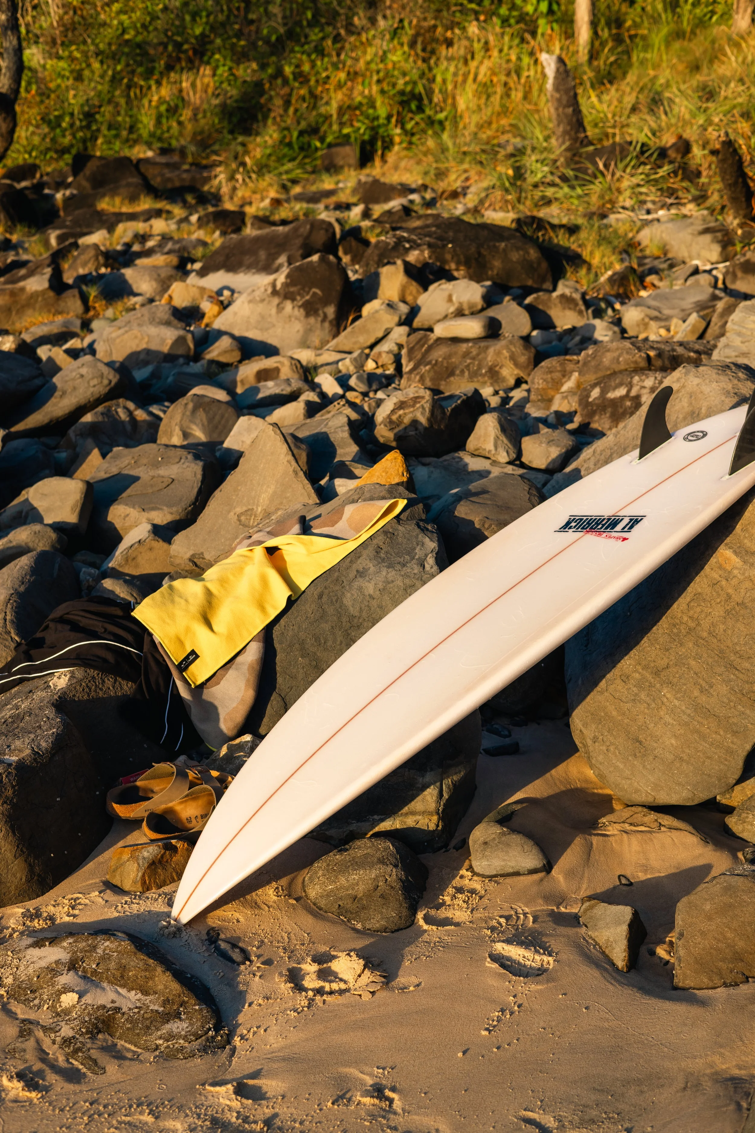 A sandy beach with rocks, a white surfboard, a yellow towel, and flip flops laid on the sand near the rocks, with grassy vegetation in the background.