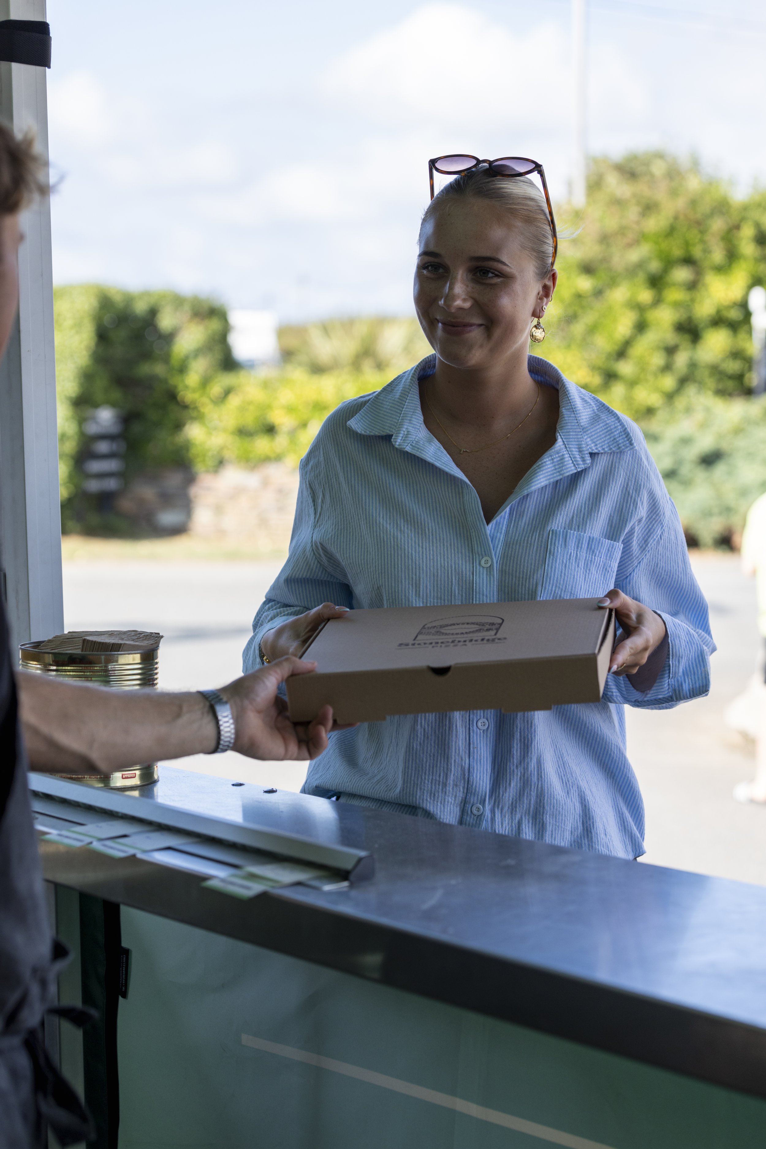 A young woman with blonde hair, sunglasses on her head, and wearing a light blue striped shirt, is smiling as she receives a pizza box from someone whose arm is visible. They are standing outdoors near a green counter with cans of food and papers on 