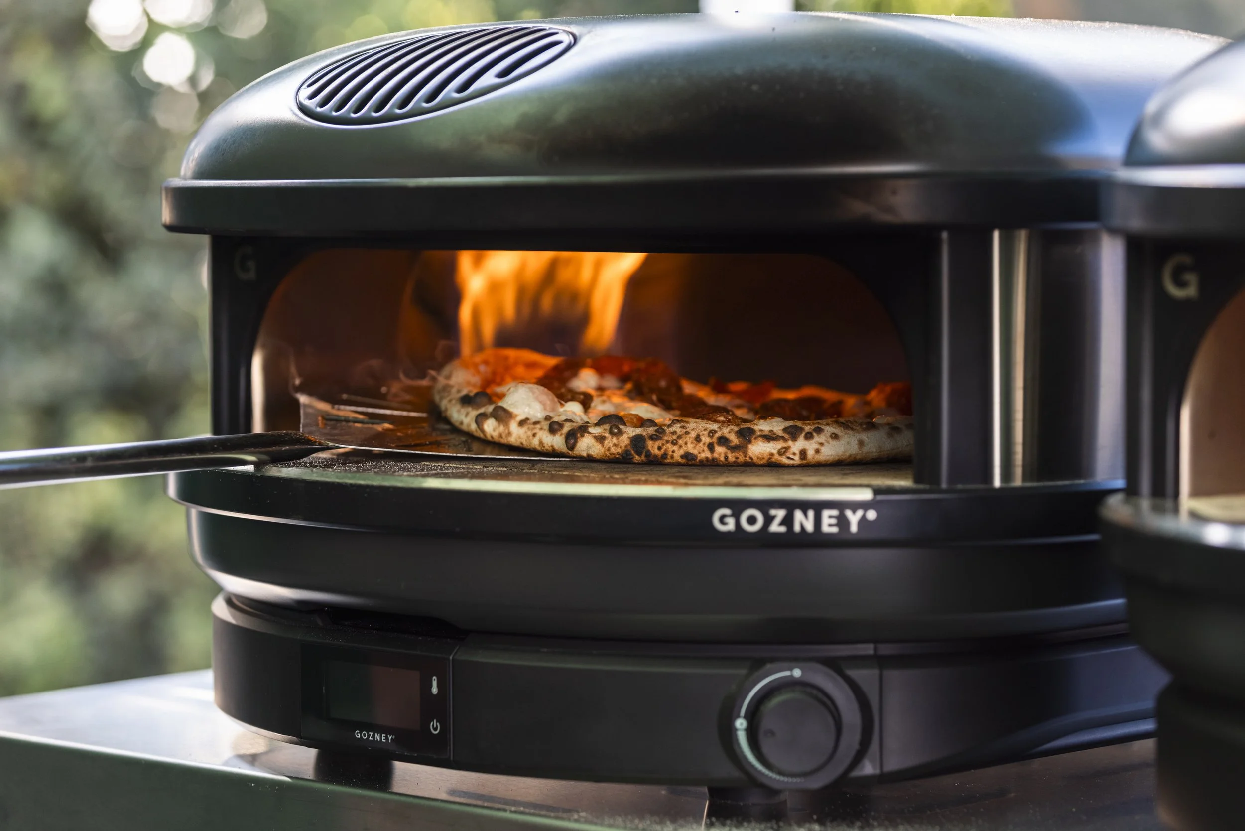 A portable outdoor pizza oven with a flame on a pizza inside, branded Gozney, sitting on a gray surface with blurred greenery in background.