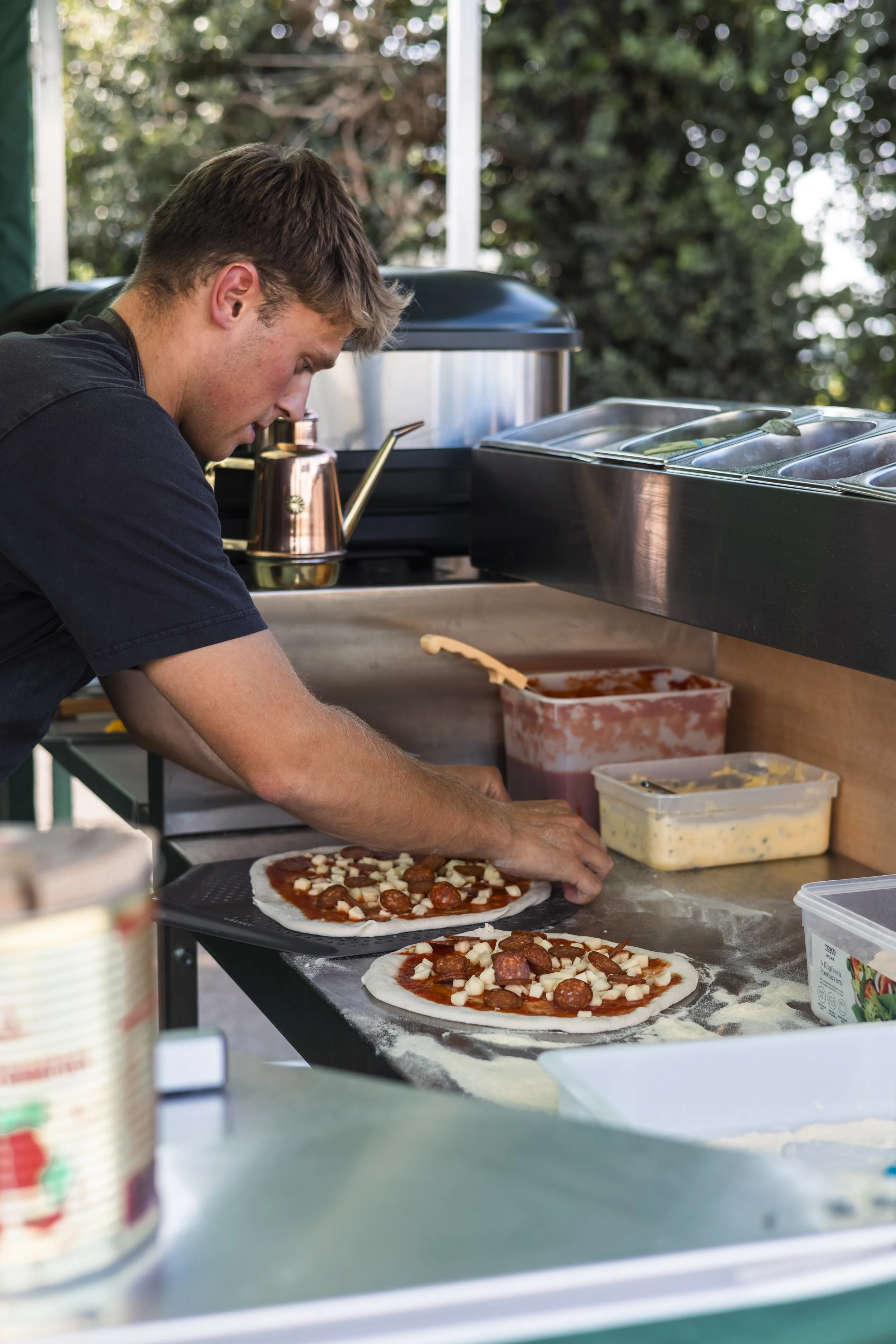 A young man makes pizzas at an outdoor food stand, spreading tomato sauce on pizza dough with toppings like pepperoni and cheese.