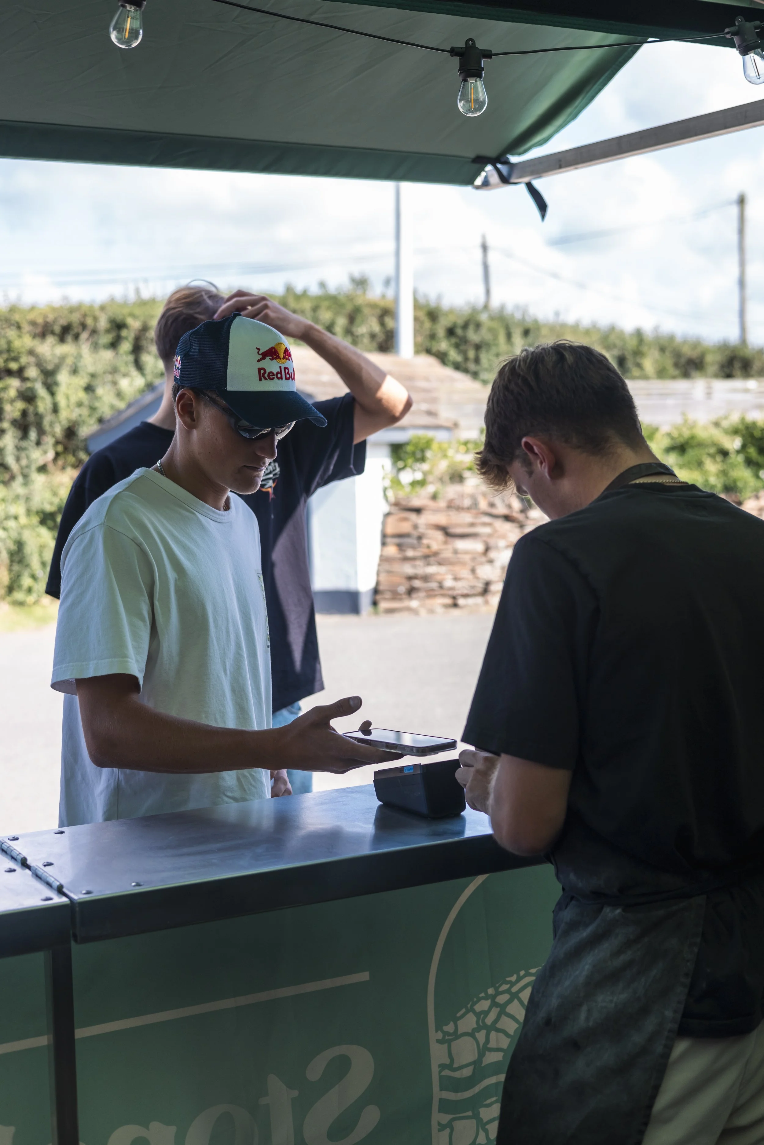 Three young men are standing at a table under a canopy, with two of them looking at a smartphone and the third touching his head. They are outdoors, with a background of trees, a building, and power lines. One man is wearing sunglasses and a Red Bull