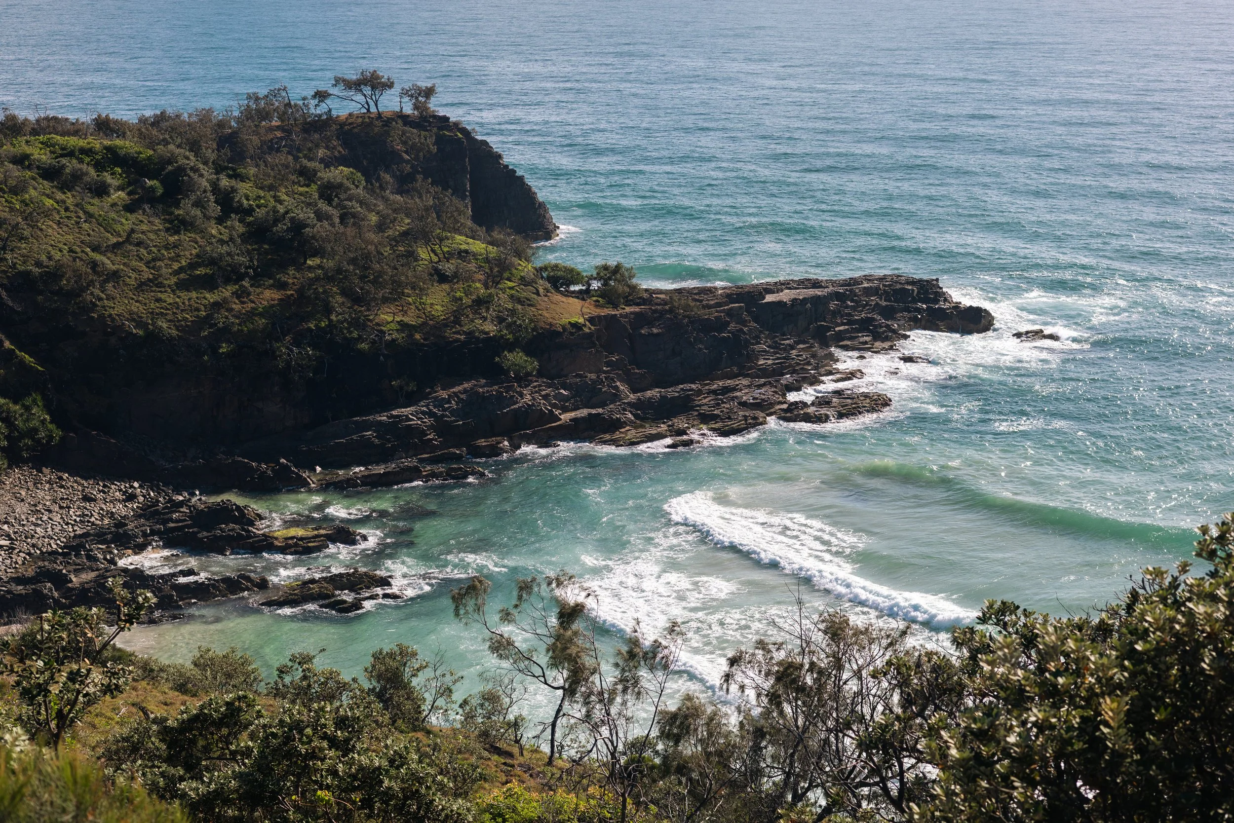 Cliffside overlooking the ocean with green vegetation and rocky shoreline, calm blue water, and small waves.