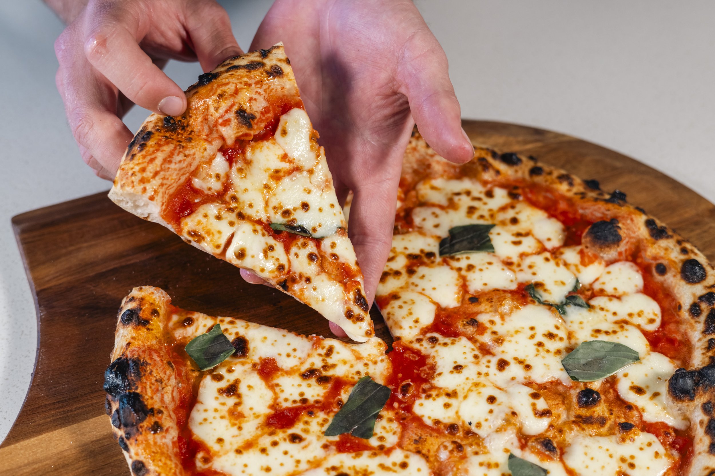 Close-up of a hand picking up a slice of cheese pizza with basil leaves on top, with remaining pizza slices on a wooden board.