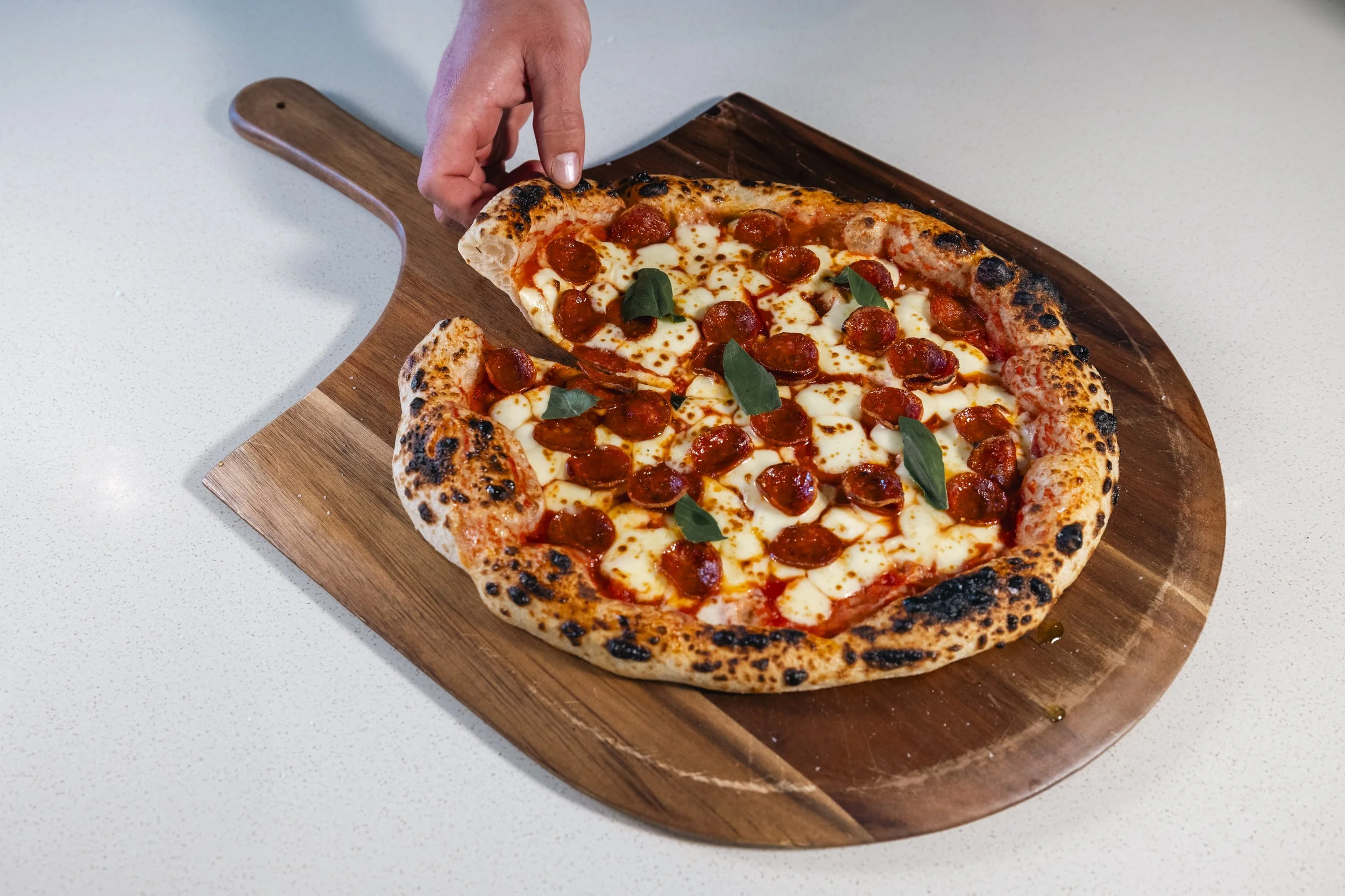 Close-up of a pepperoni pizza on a wooden serving board, with a hand picking up a slice. The pizza has a crust with charred spots, melted cheese, pepperoni slices, and basil leaves on top. The background is a white countertop.