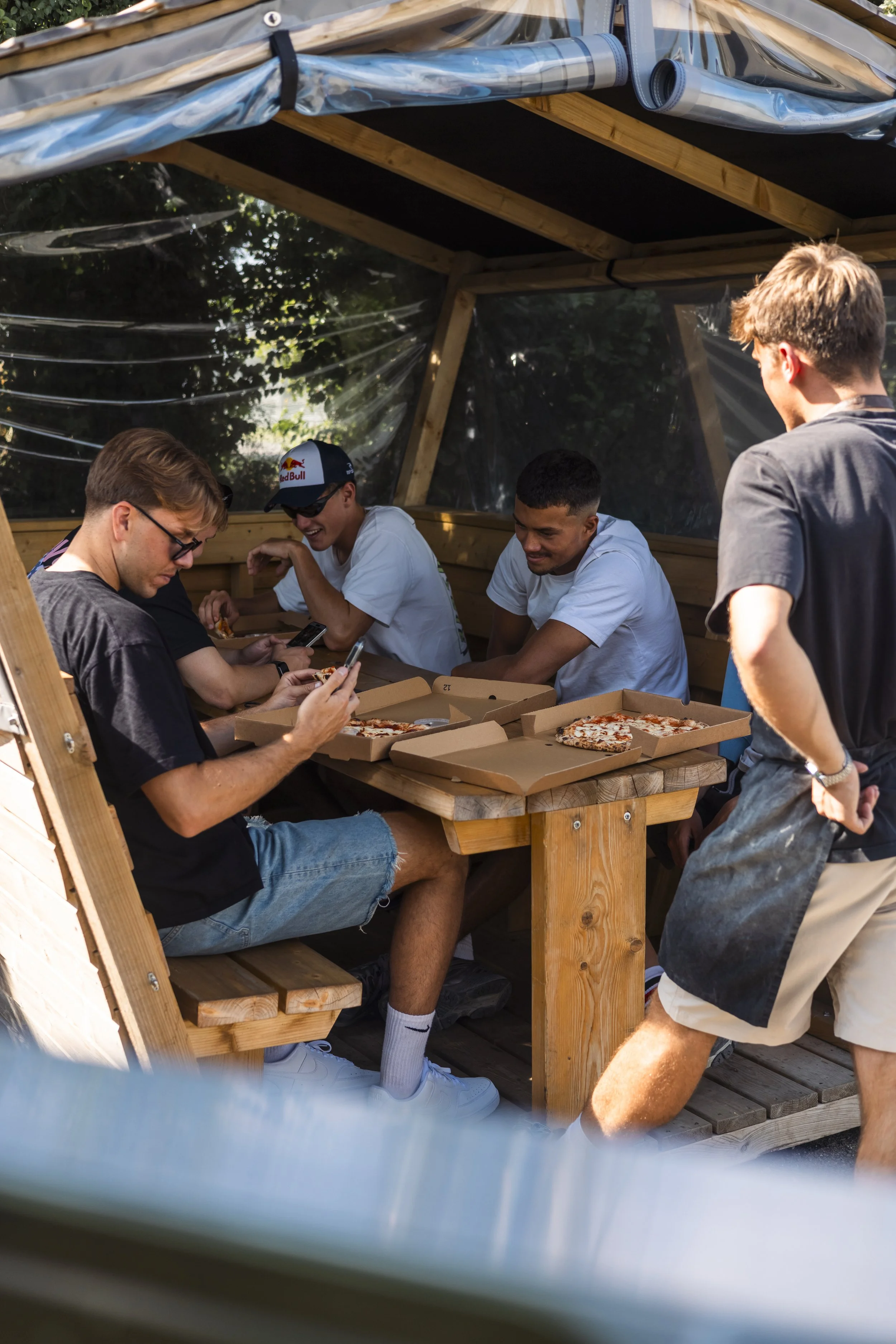 Young men sitting at a wooden picnic table with pizza boxes, some looking at their phones, under a makeshift outdoor shelter.