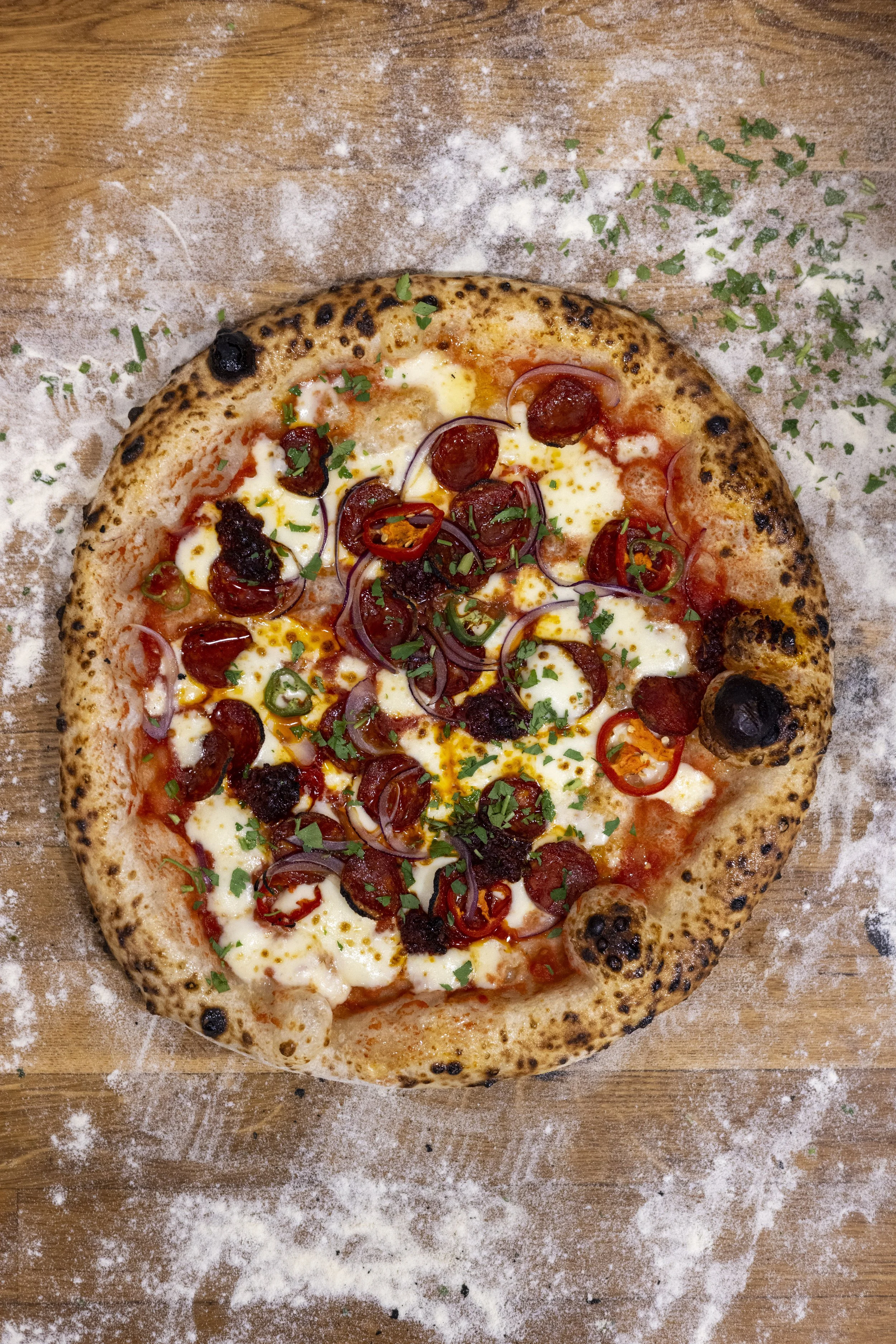 Pizza with pepperoni, red onions, green peppers, cheese, and herbs on a wooden surface with flour scattered.