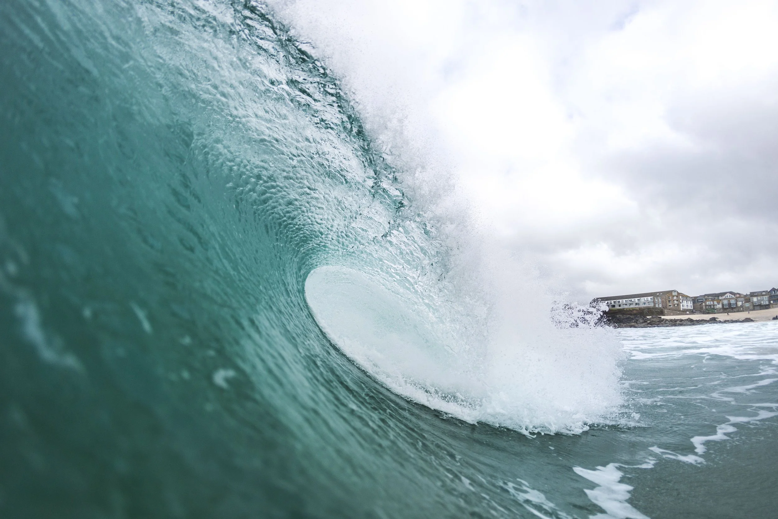 A large ocean wave about to break near a shoreline with buildings in the background under an overcast sky.