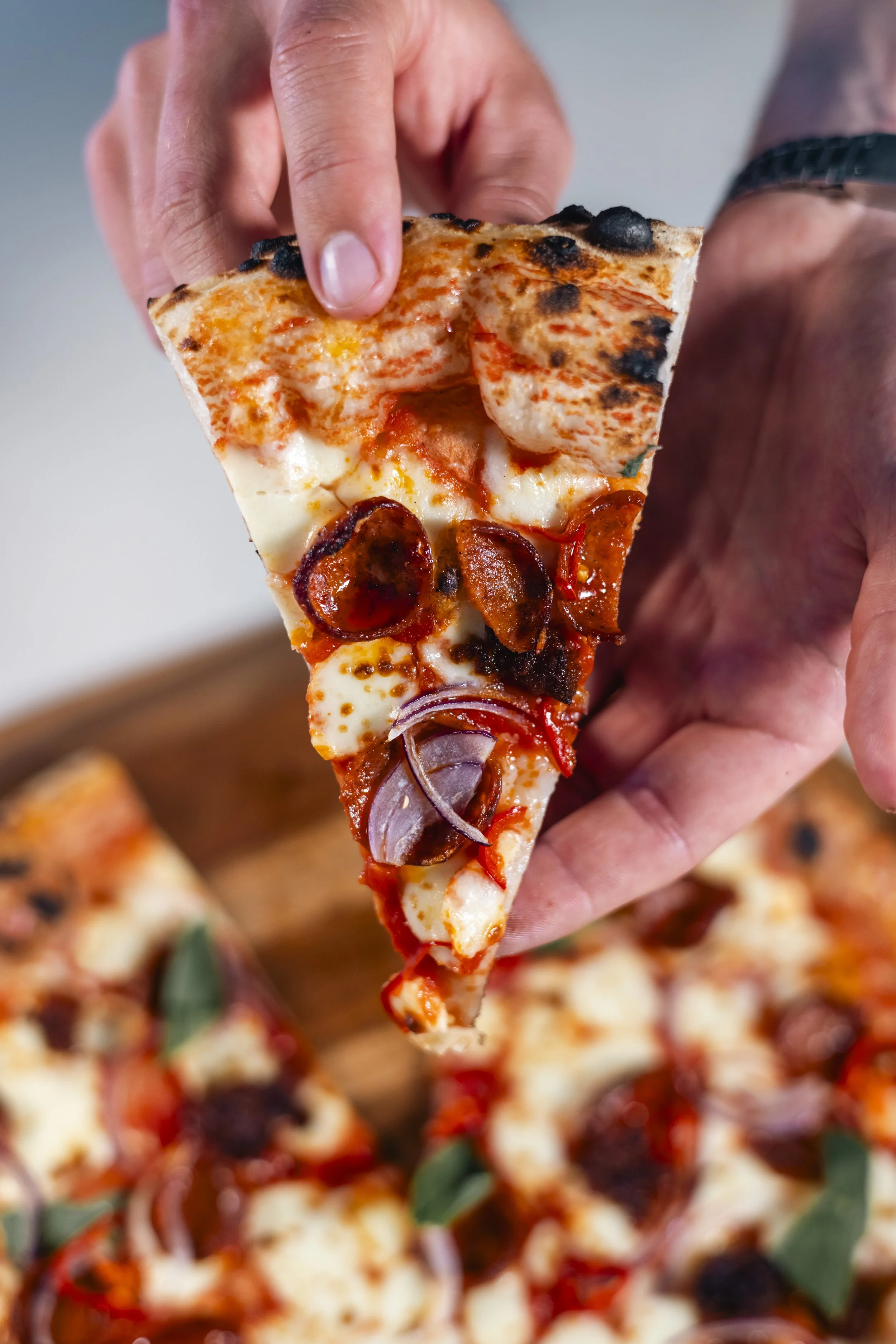 Close-up of a hand holding a slice of pepperoni pizza with melted cheese, tomato sauce, blackened spots from charring, and onions, with a blurred pizza in the background.