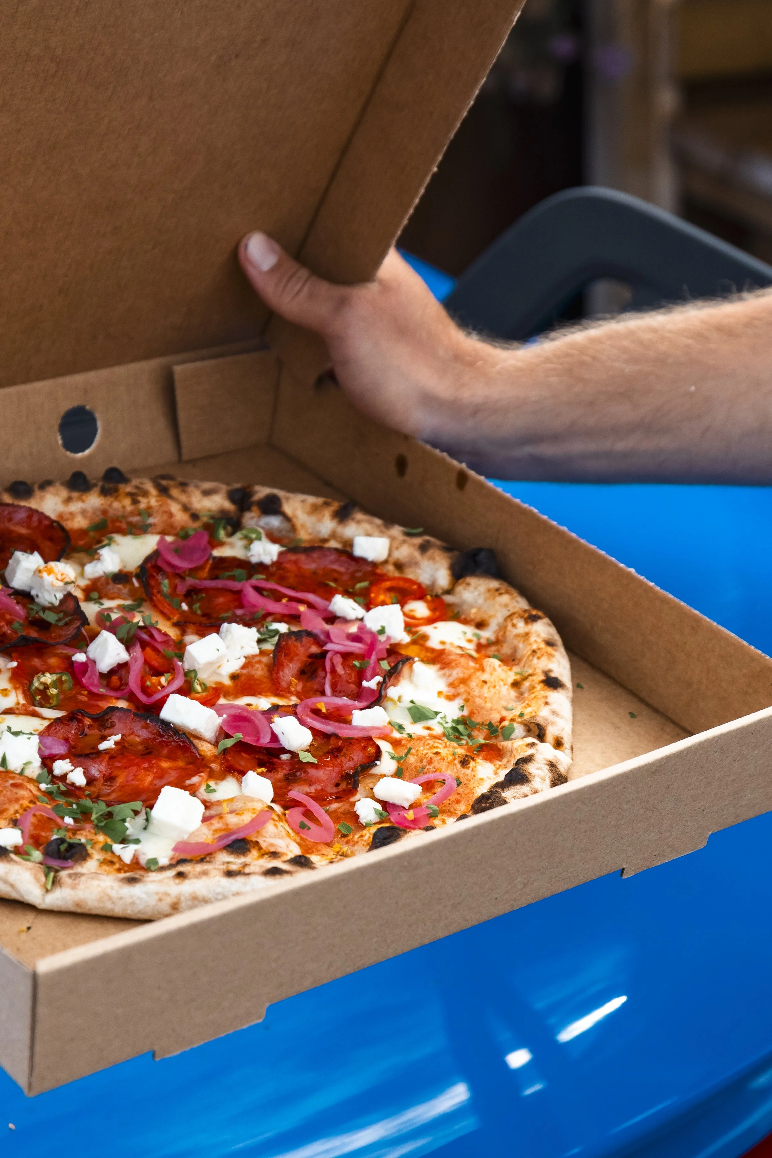 Close-up of a pepperoni pizza with toppings in an open cardboard pizza box on a blue table.