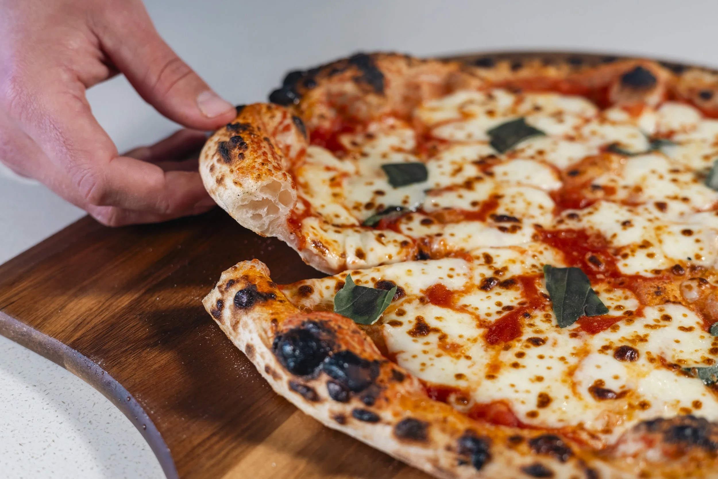 Close-up of a hand pulling a slice of cheese pizza with a thick crust, melted mozzarella cheese, tomato sauce, and basil leaves on a wooden serving board.