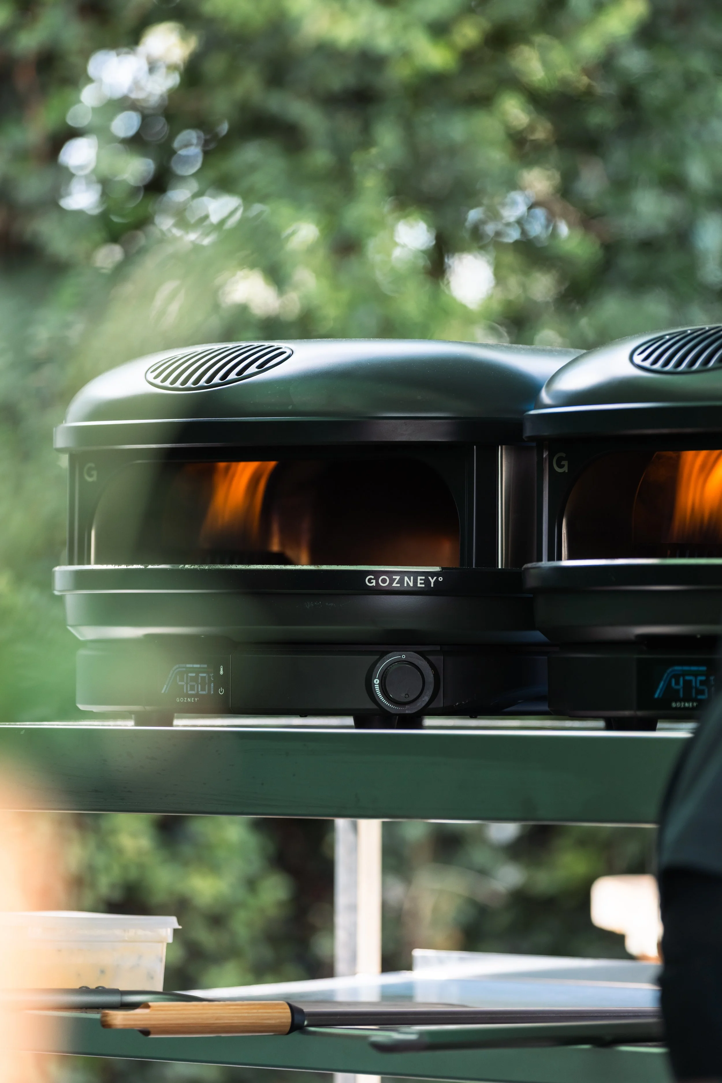 Close-up of a Gozney pizza oven with a blurred green outdoor background.