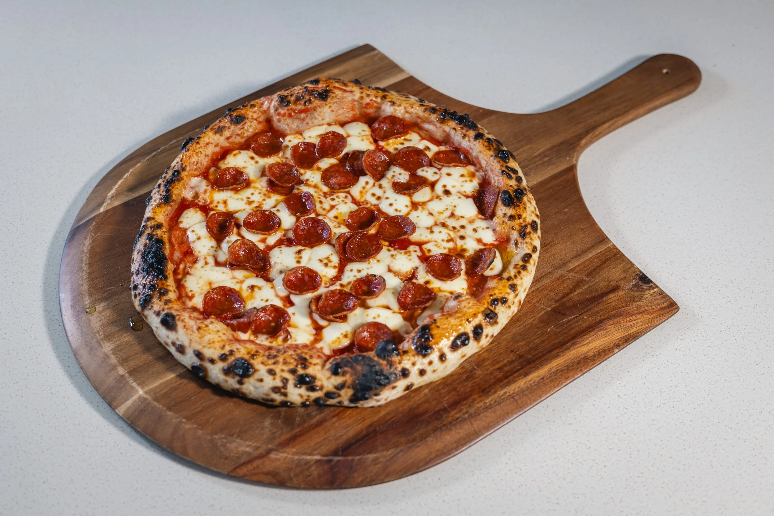 A pepperoni pizza with melted cheese on a wooden pizza peel, placed on a white countertop.