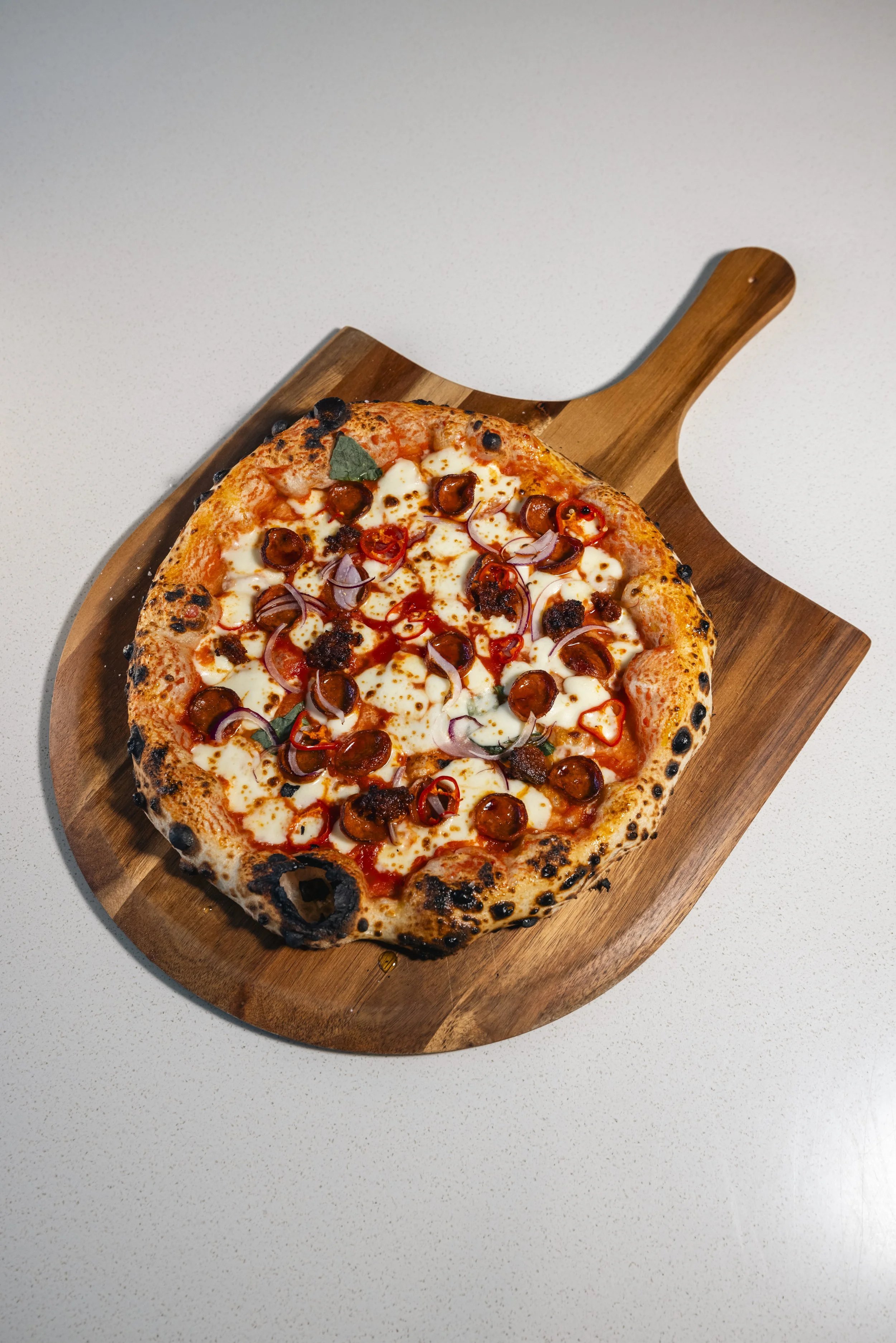 A pizza with cheese, pepperoni, red onions, and basil on a wooden pizza peel.