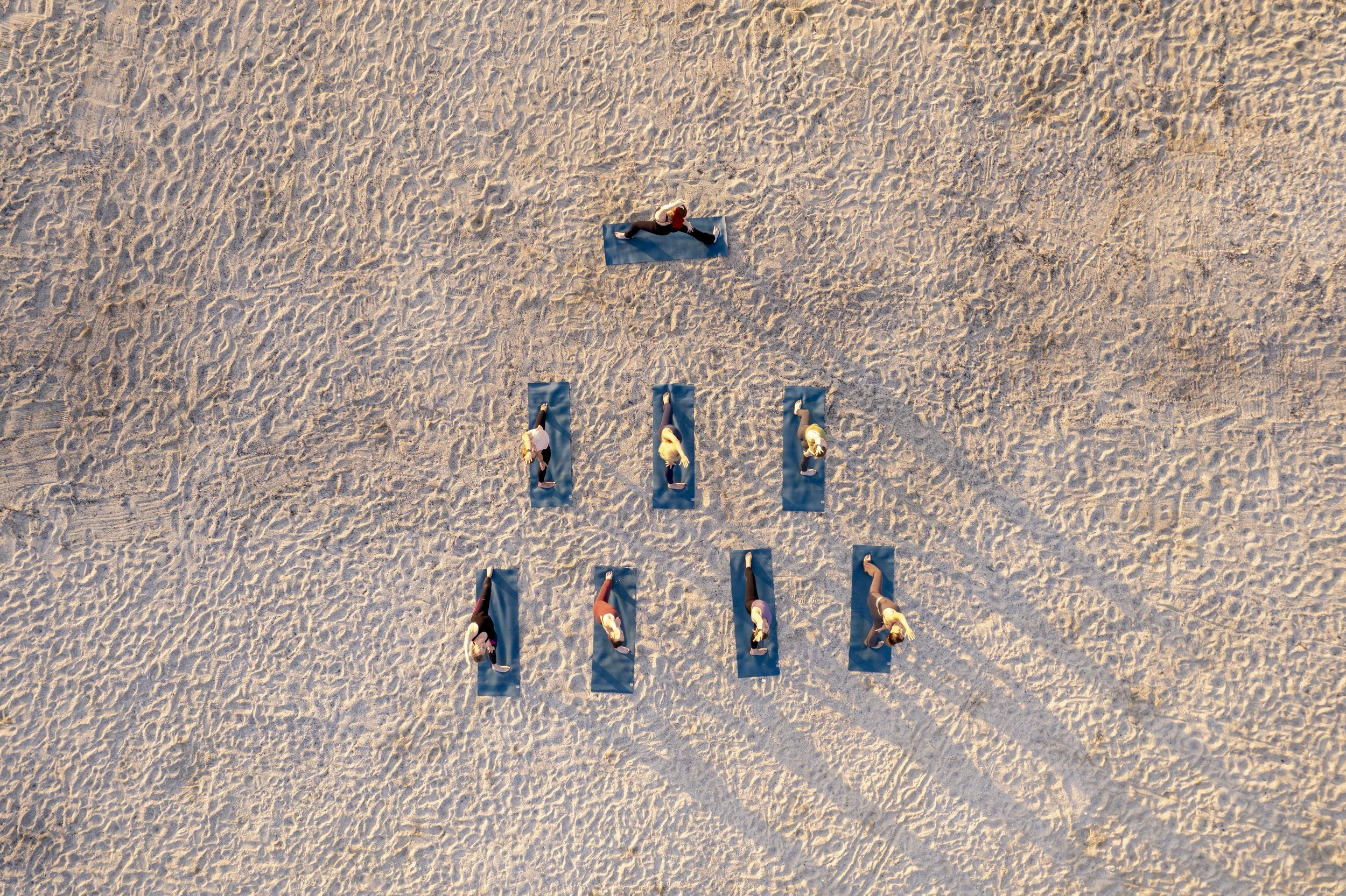 An aerial view of people practicing yoga on blue mats on sandy beach, spaced evenly apart.