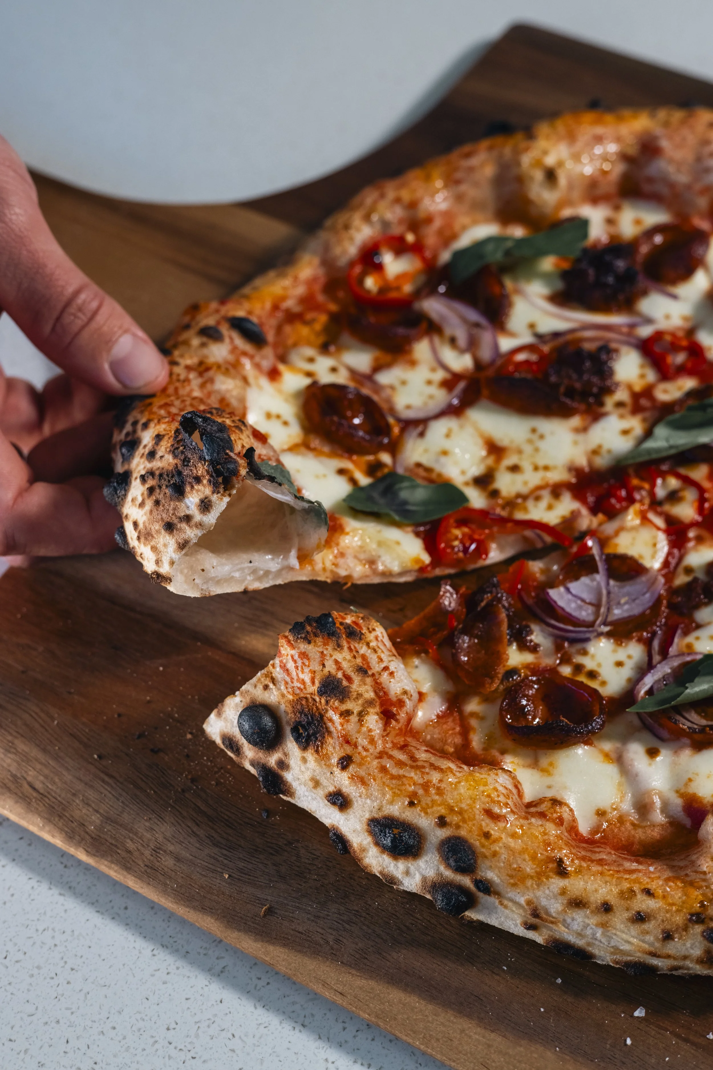 Close-up of a pizza with pepperoni, red onions, basil leaves, and a cheese topping, held by a person near its crust, on a wooden serving board.