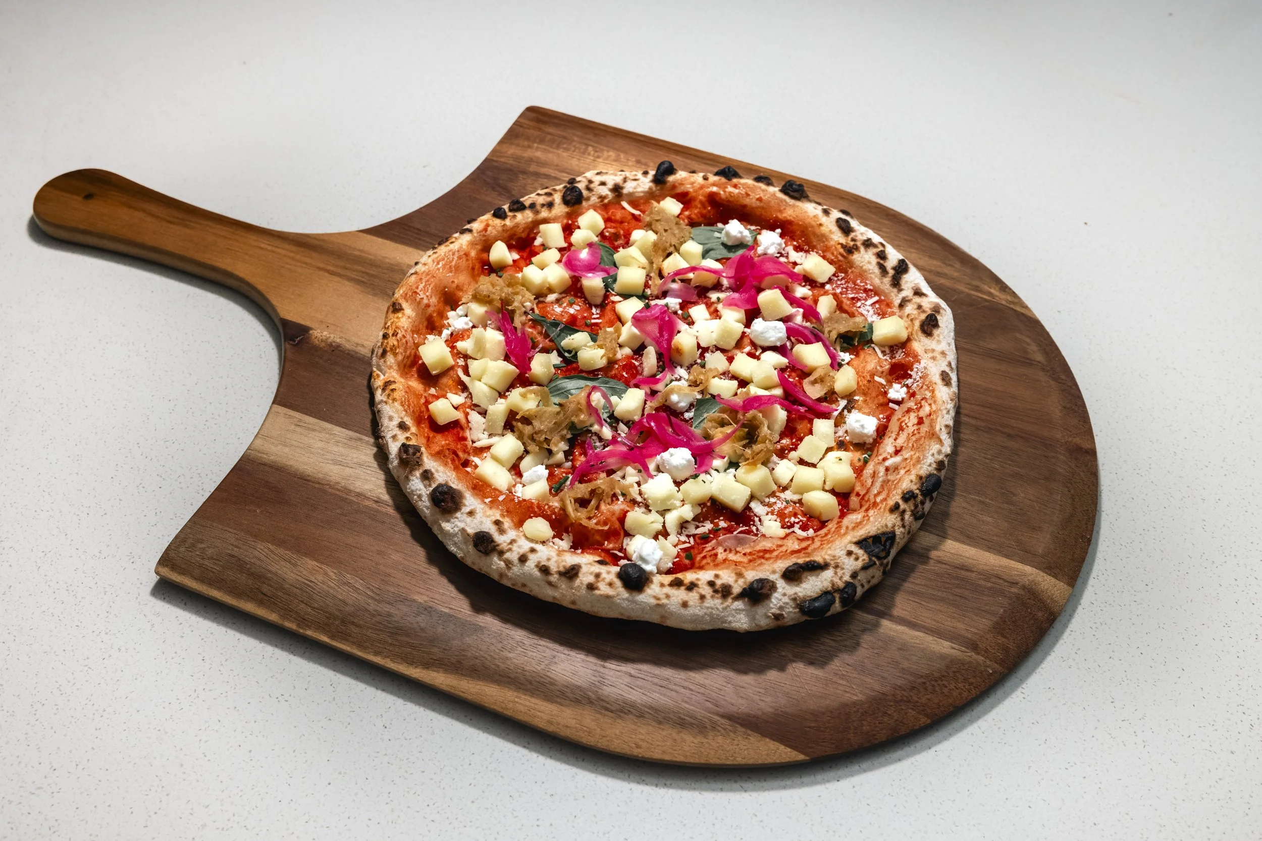 A pizza with cheese, pickled vegetables, and herbs on a wooden serving board.