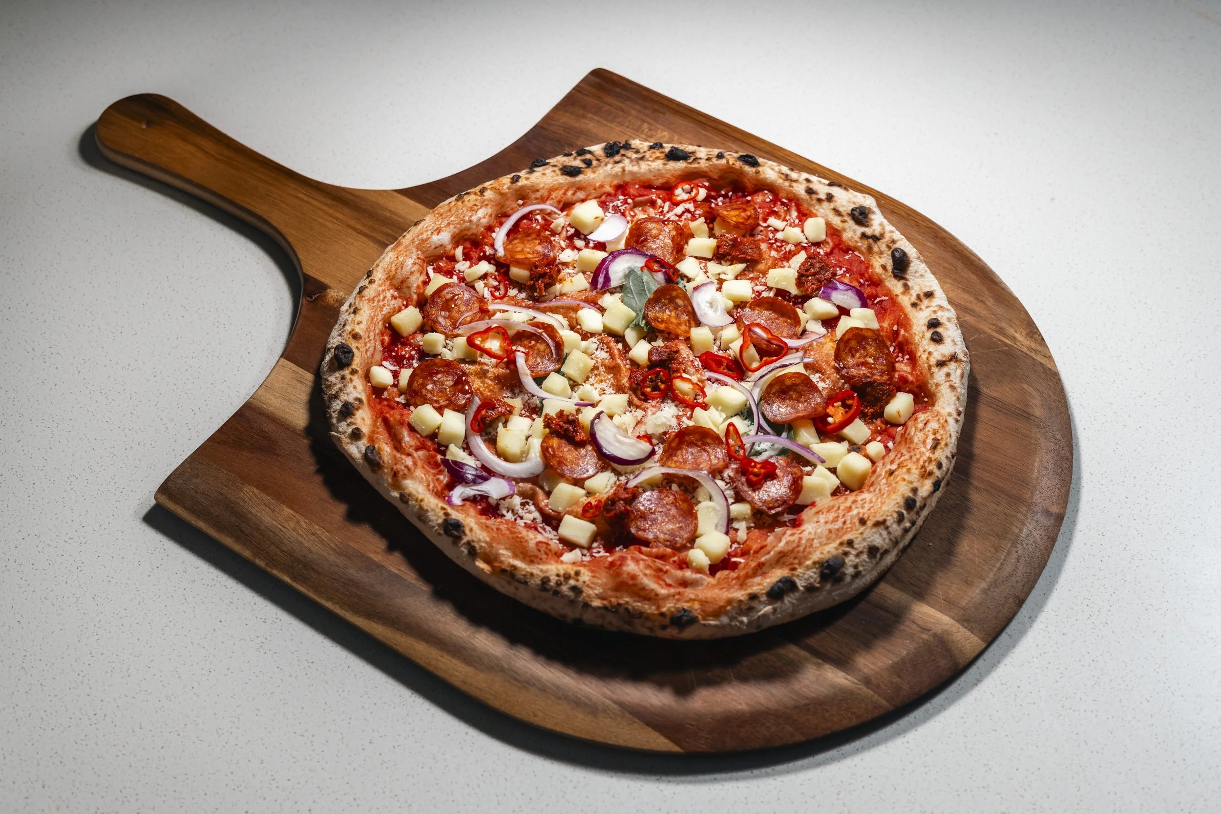 Pizza with pepperoni, cheese, red onion, and chili peppers on a wooden pizza peel.