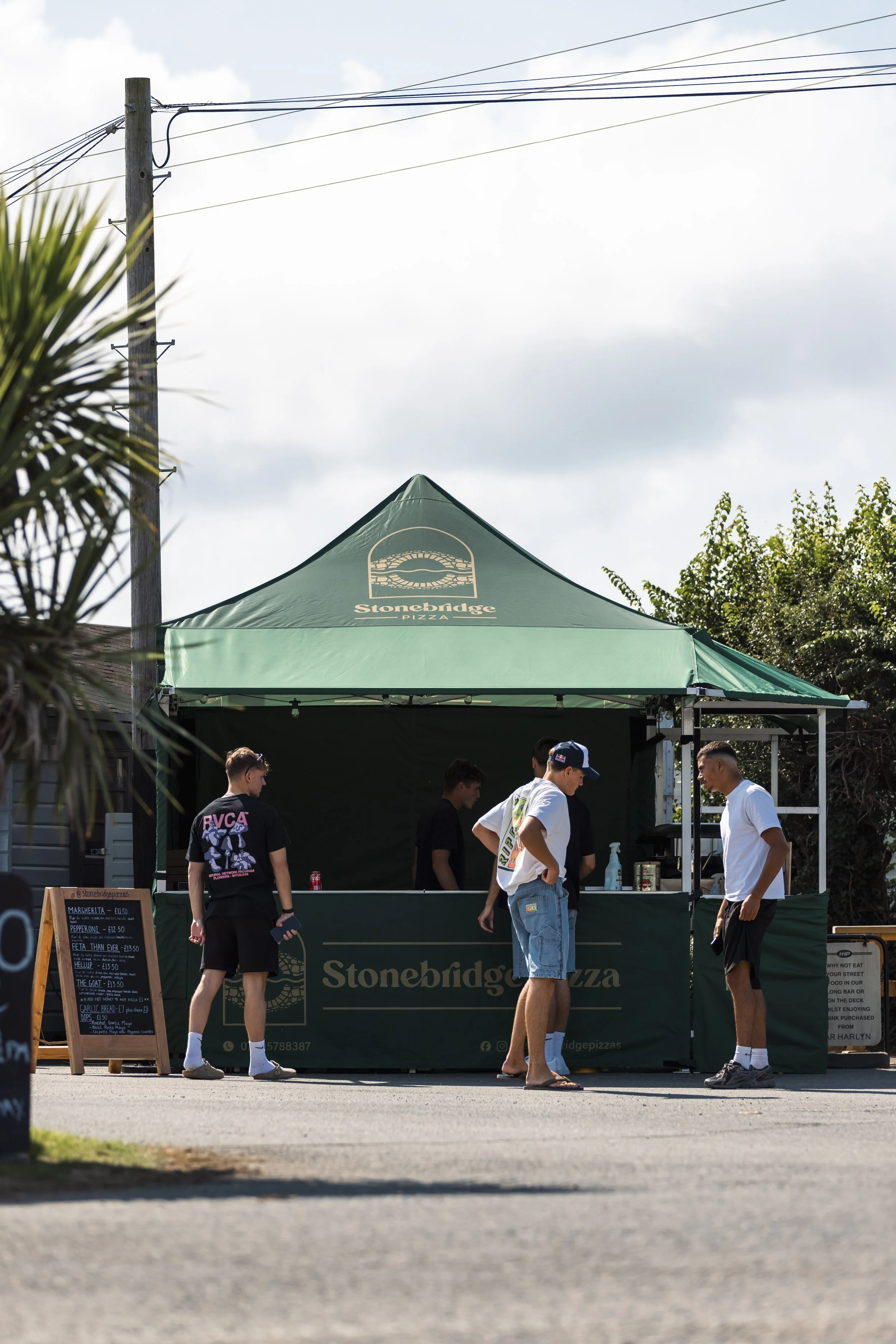 People standing and talking in front of a green pizza stand with a Stonebridge Pizza logo on top, outdoor setting with trees and a pole with electrical wires overhead.