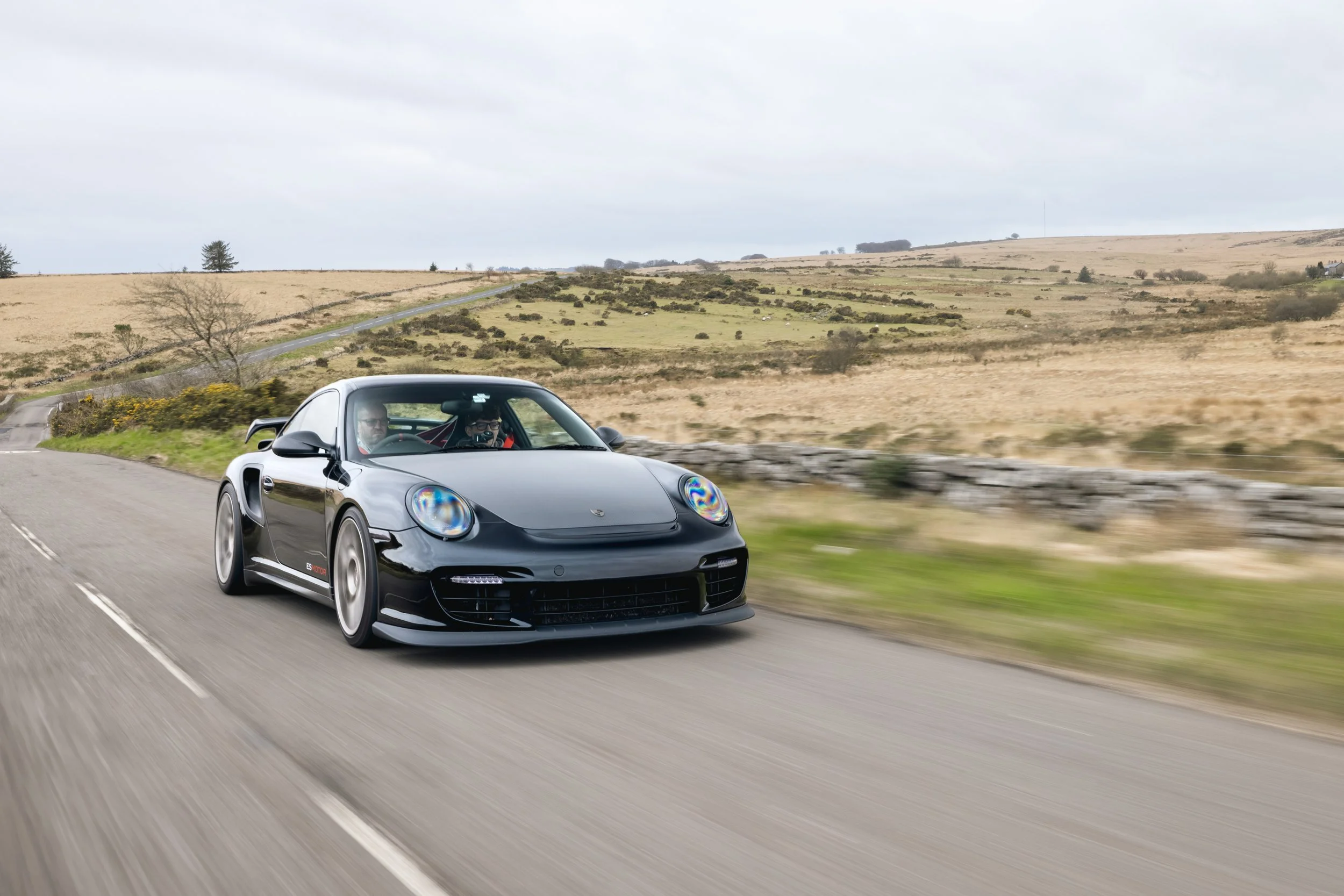 A black Porsche sports car driving on an open countryside road with two passengers visible through the windshield, in a landscape of rolling hills and sparse trees under a cloudy sky.