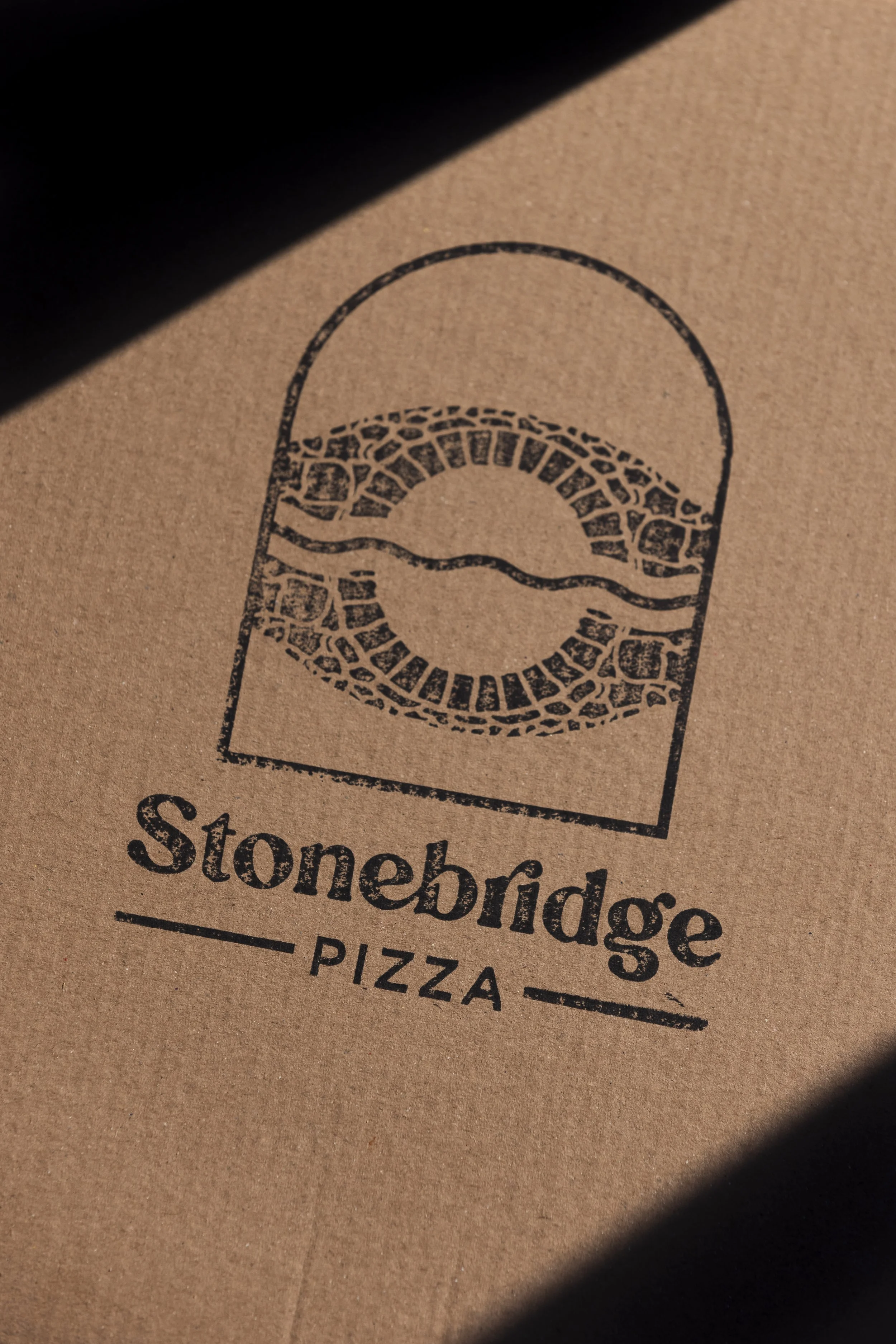 Close-up of a brown paper pizza box with a black logo and text for Stonebridge Pizzas, partially covered by shadow.
