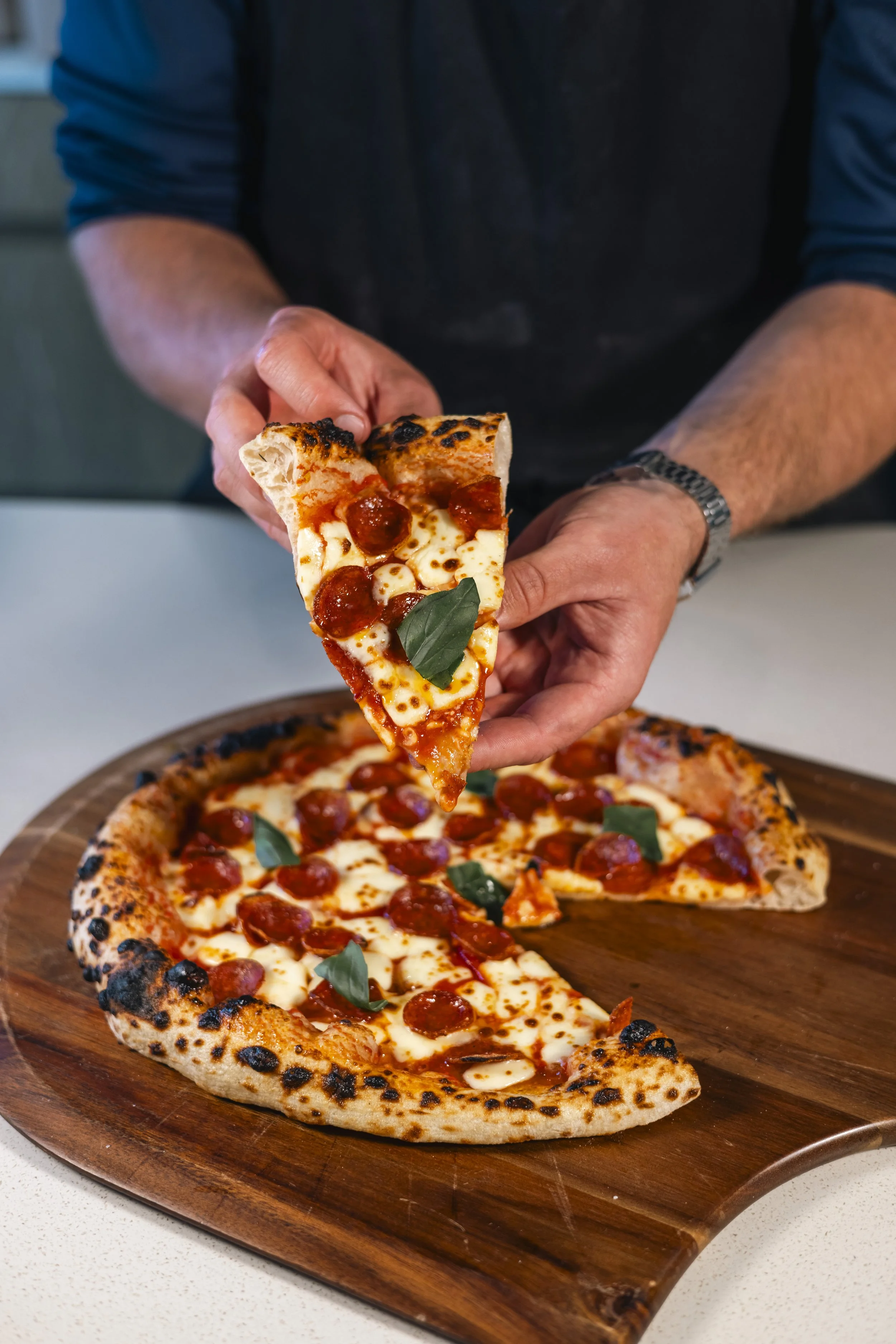 A person is holding a slice of pepperoni pizza over a wooden pizza board, with the remaining pizza visible on the board.