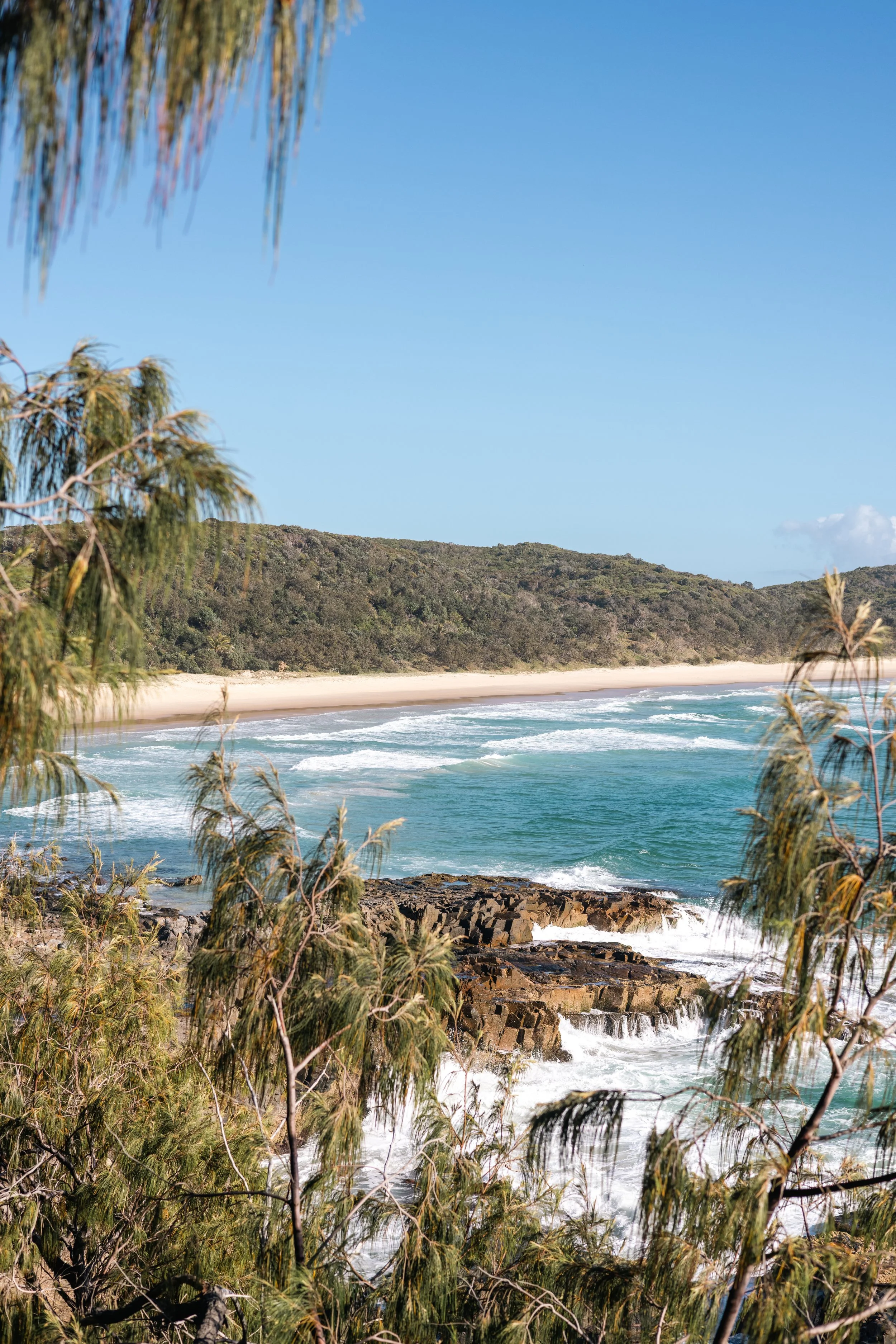Ocean waves crashing on rocky shore with sandy beach, green trees, and blue sky.