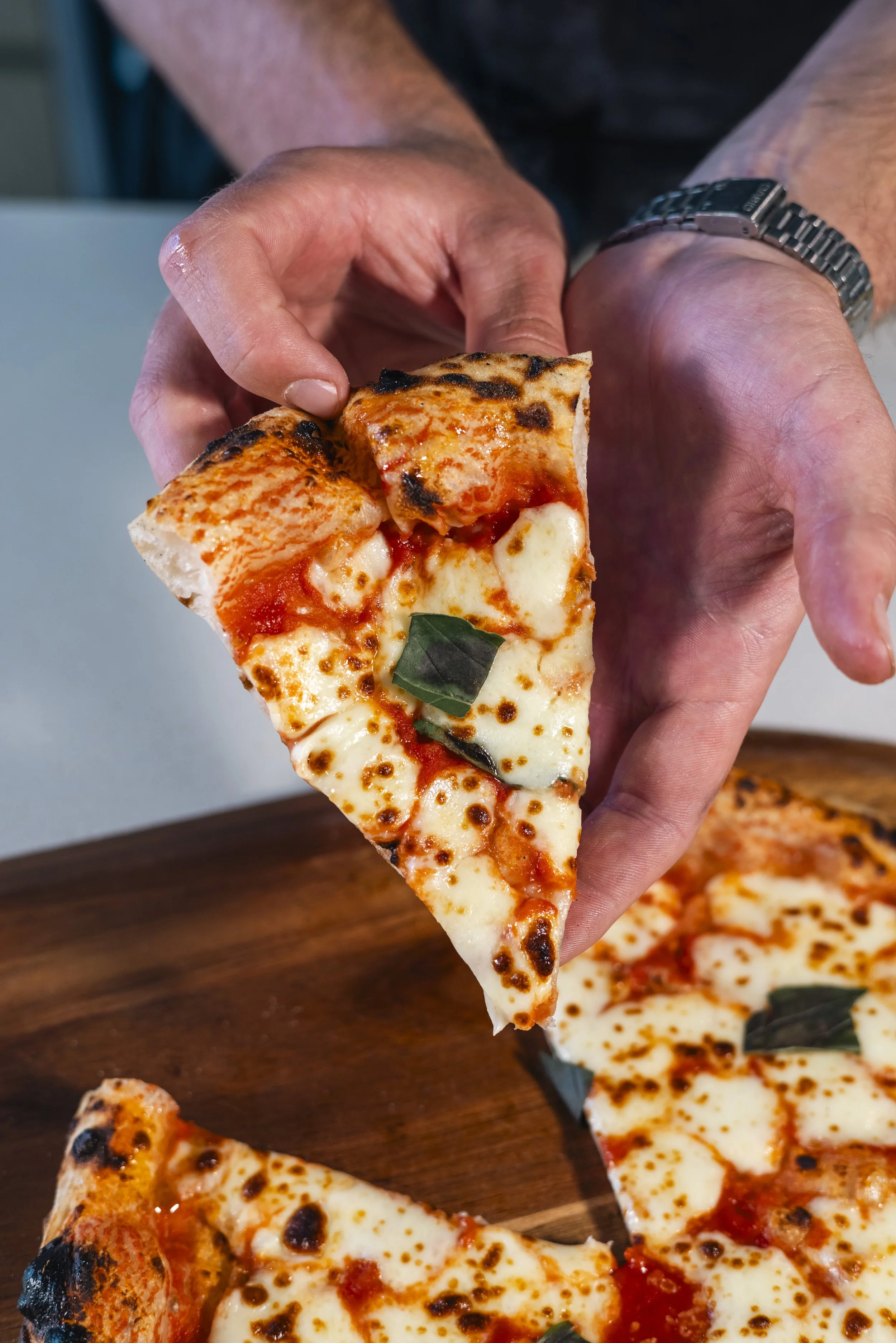 A hand holding a slice of cheese pizza with tomato sauce and basil leaf, with pizza slices on a wooden table in the background.