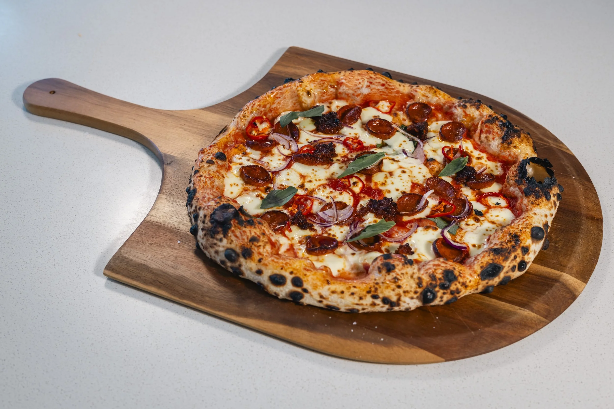 A pizza with pepperoni, red onion, and basil on a wooden serving board on a white surface.