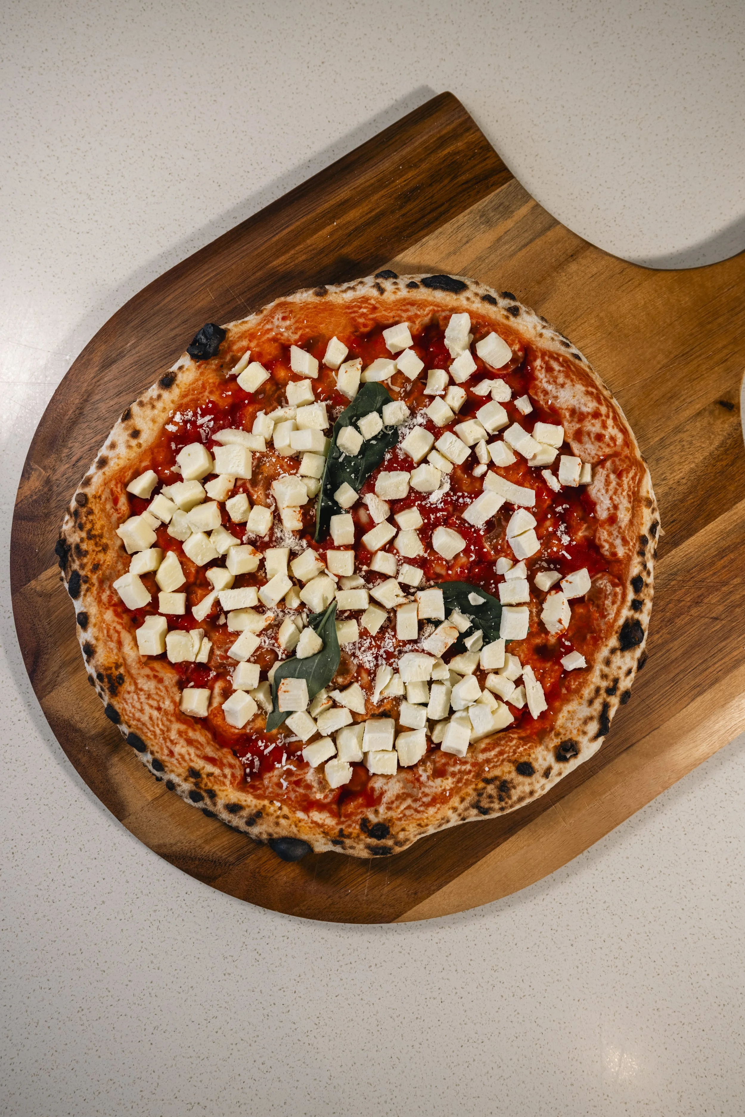 A freshly baked pizza topped with cubed mozzarella cheese, tomato sauce, and basil leaves on a round wooden pizza board.