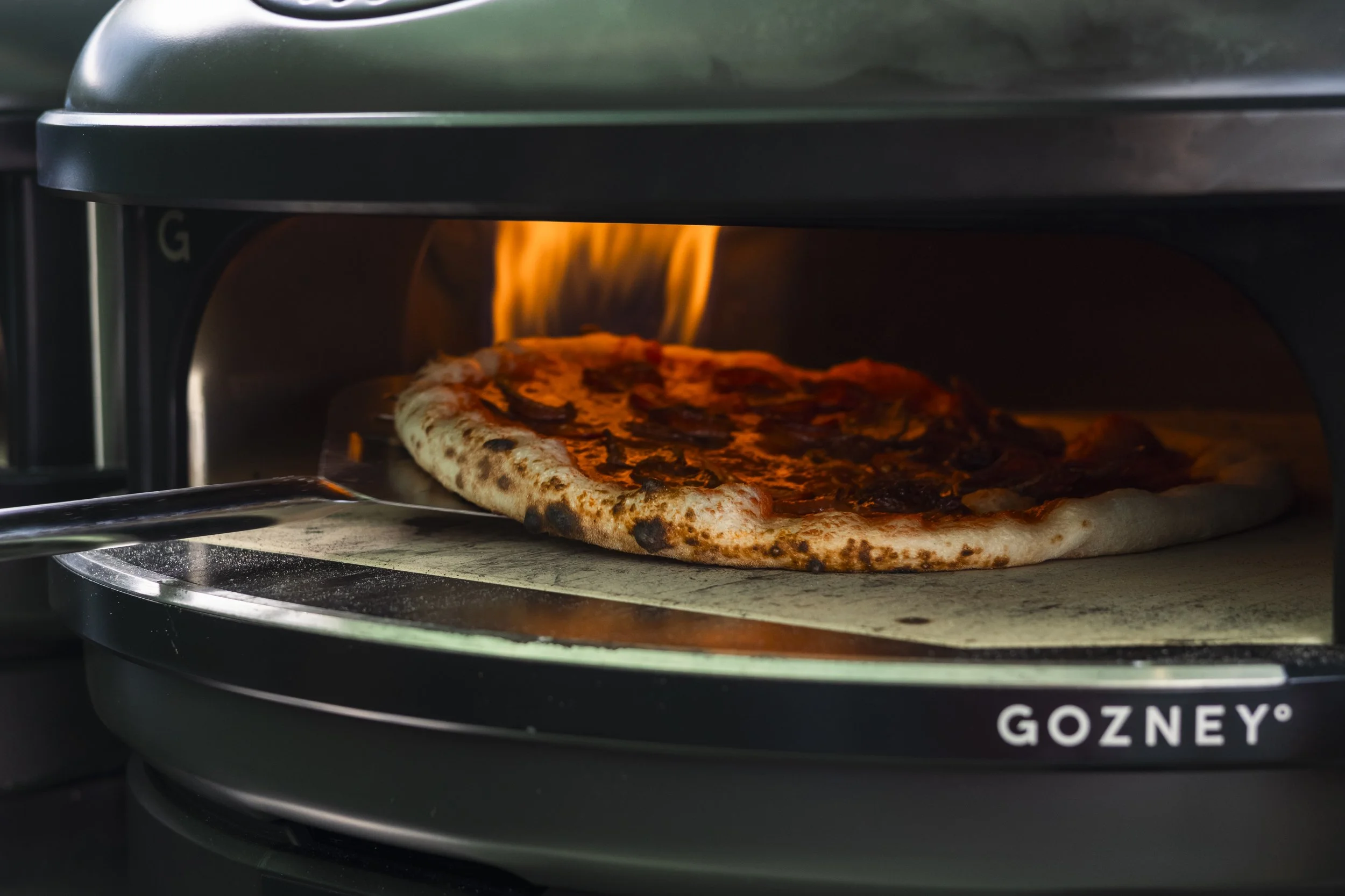 Pizza baking in a Gozney pizza oven with flames visible inside.