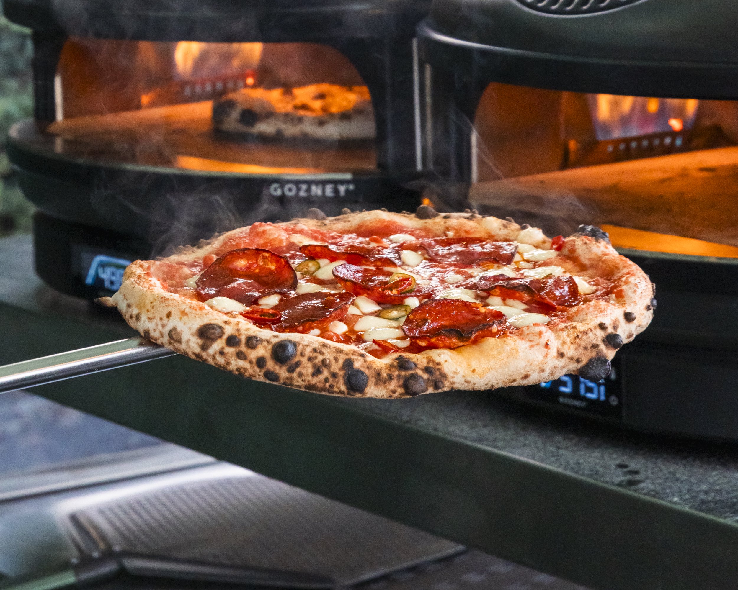 A pizza with pepperoni, sausage, and cheese baking inside a black outdoor pizza oven with flames visible in the background.