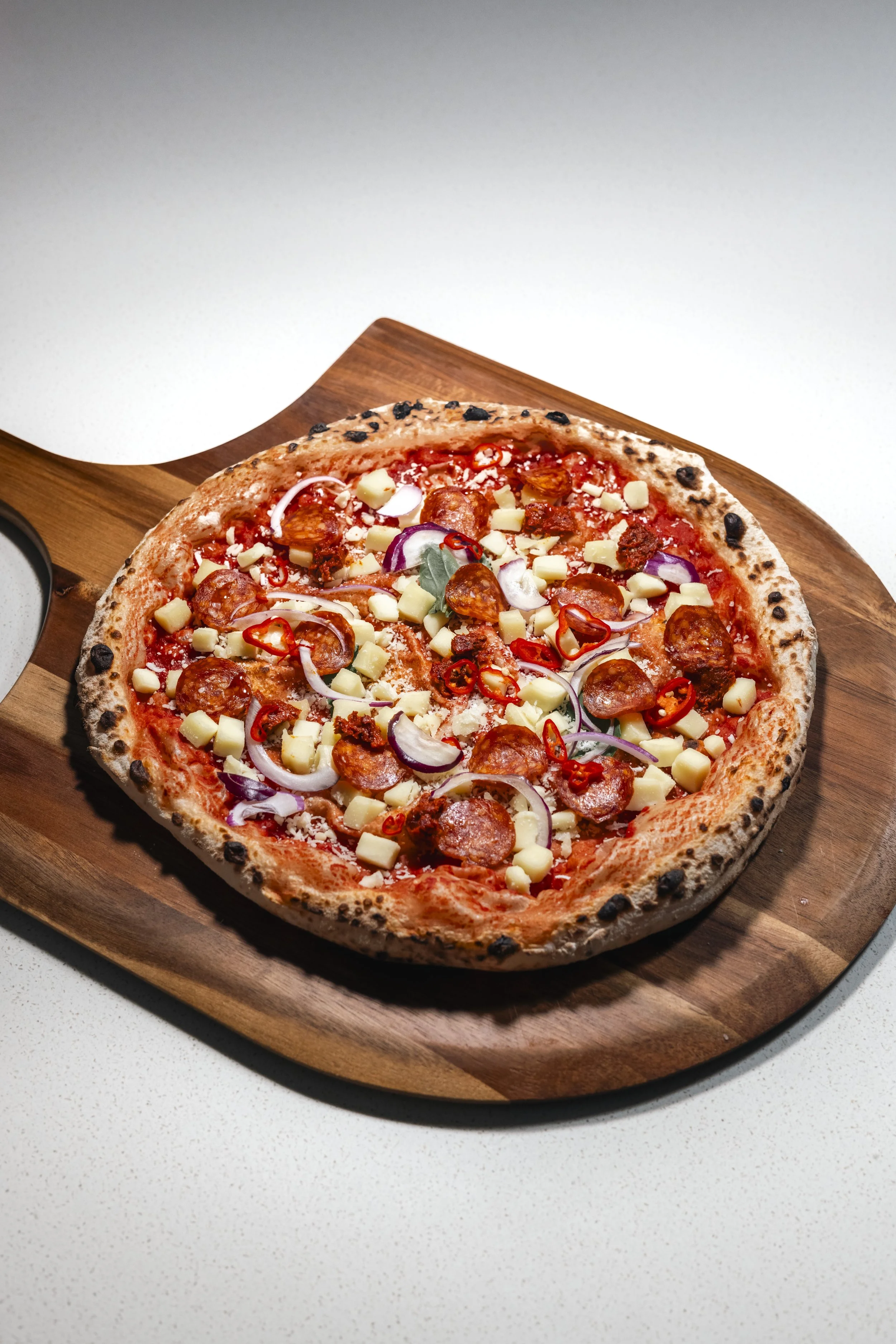 A pizza with pepperoni, cheese, red onions, red chili slices, and green leaves on a wooden serving board.
