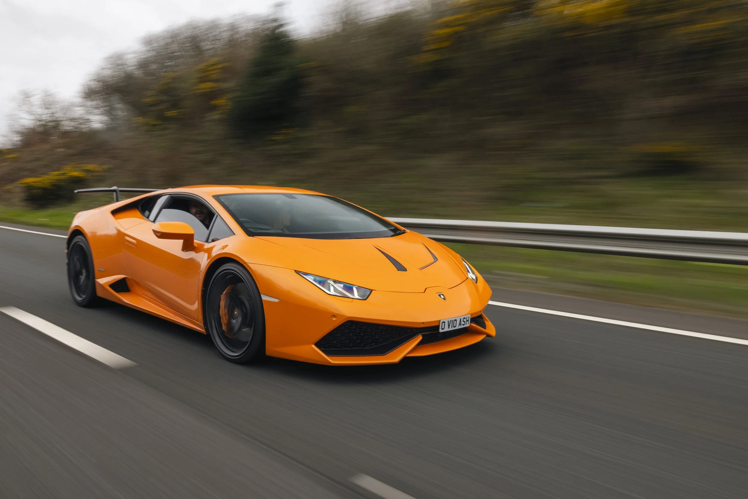An orange Lamborghini sports car driving on a highway with a blurred background of trees and overcast sky.