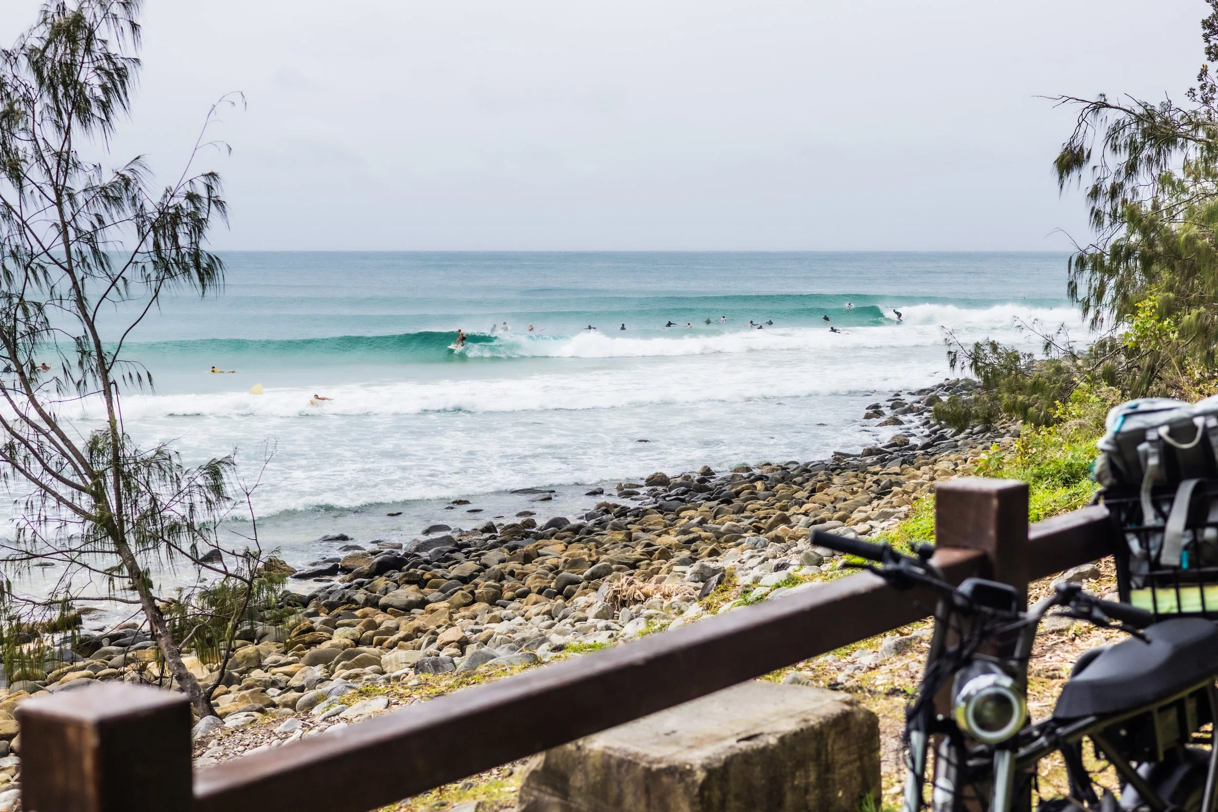 A view of a rocky shoreline with trees and a bench with a bicycle, overlooking the ocean where multiple surfers are riding waves.
