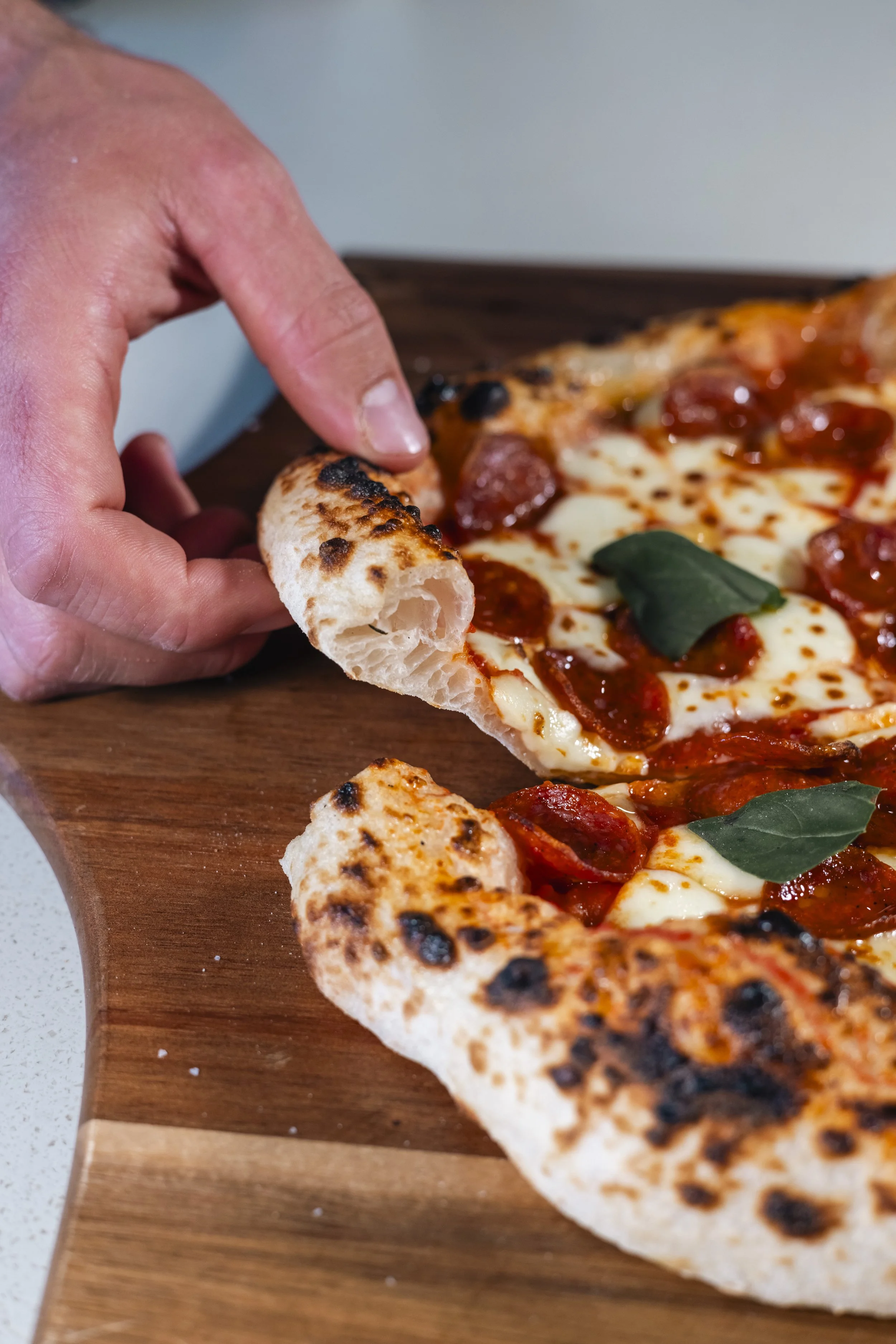 Close-up of a hand removing a slice of pepperoni pizza from a wooden serving board. The pizza has a golden crust with some charred spots, melted cheese, pepperoni slices, and fresh basil leaves.