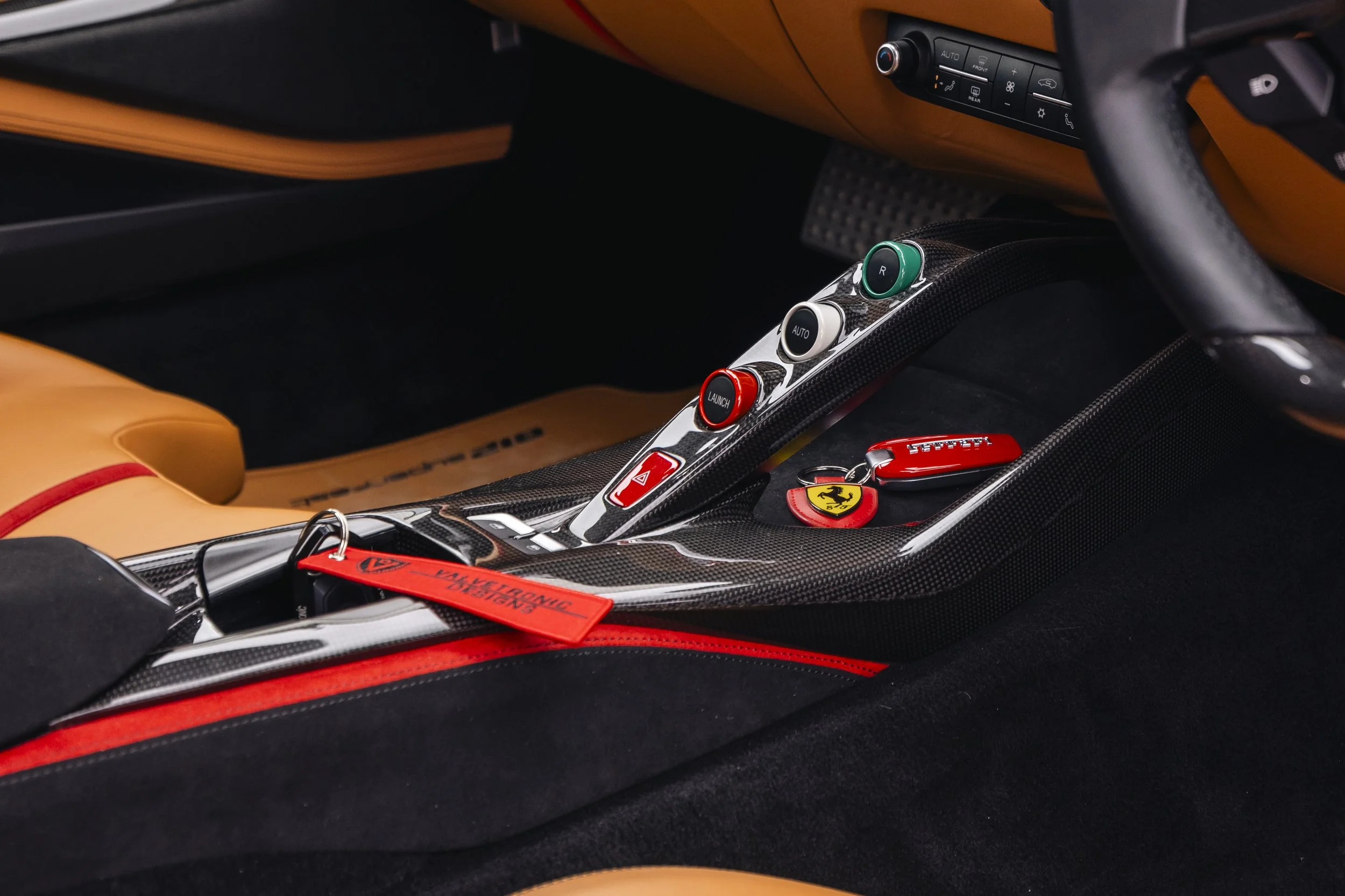 Close-up of a Ferrari FXX track car interior showing a carbon fiber center console with red and green buttons, Ferrari logo keychain, and tan leather interior.