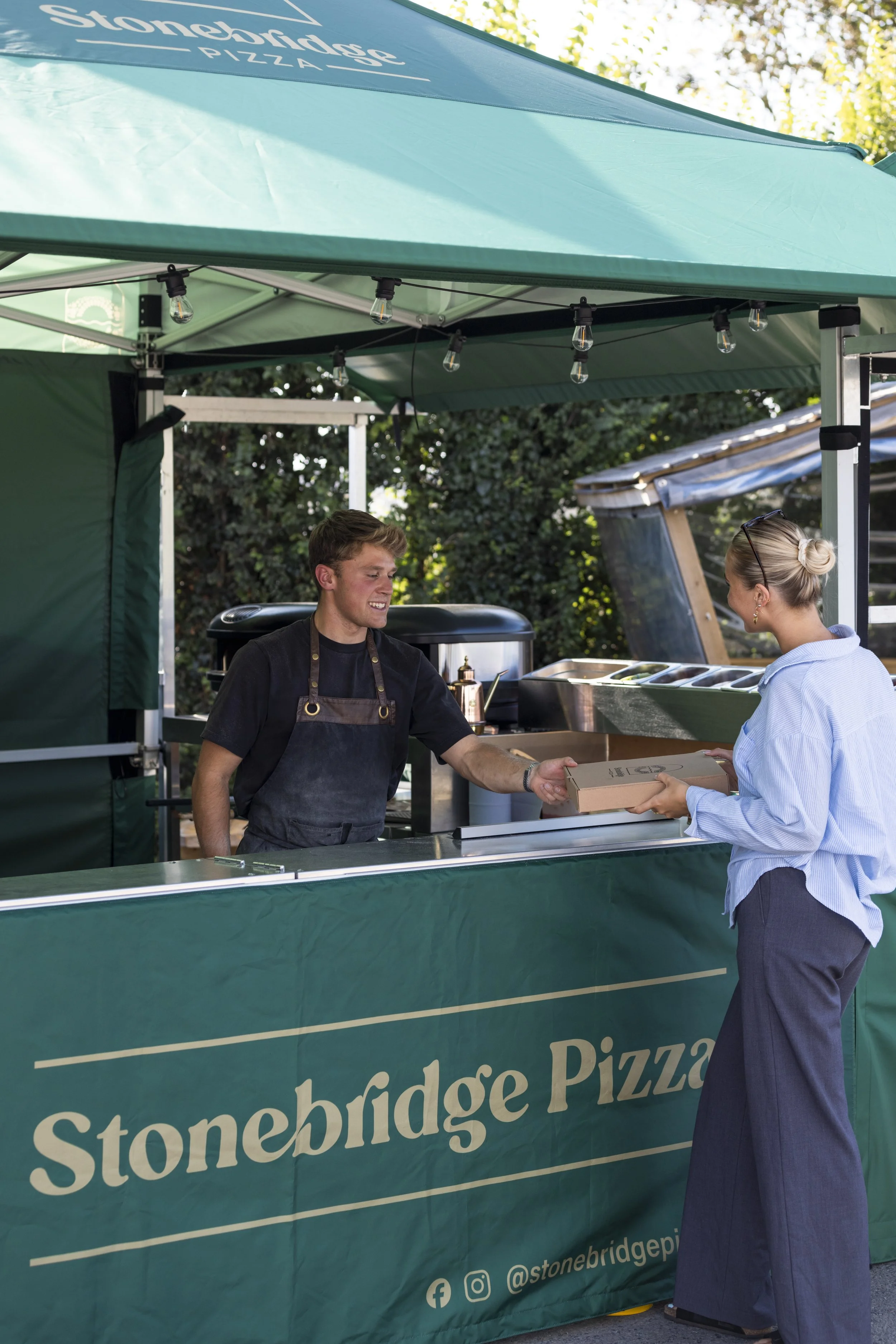 A man and a woman exchanging a pizza box at a pizza stand under a green canopy with the text 'Stonebridge Pizza' on it, outdoor setting with trees in the background.