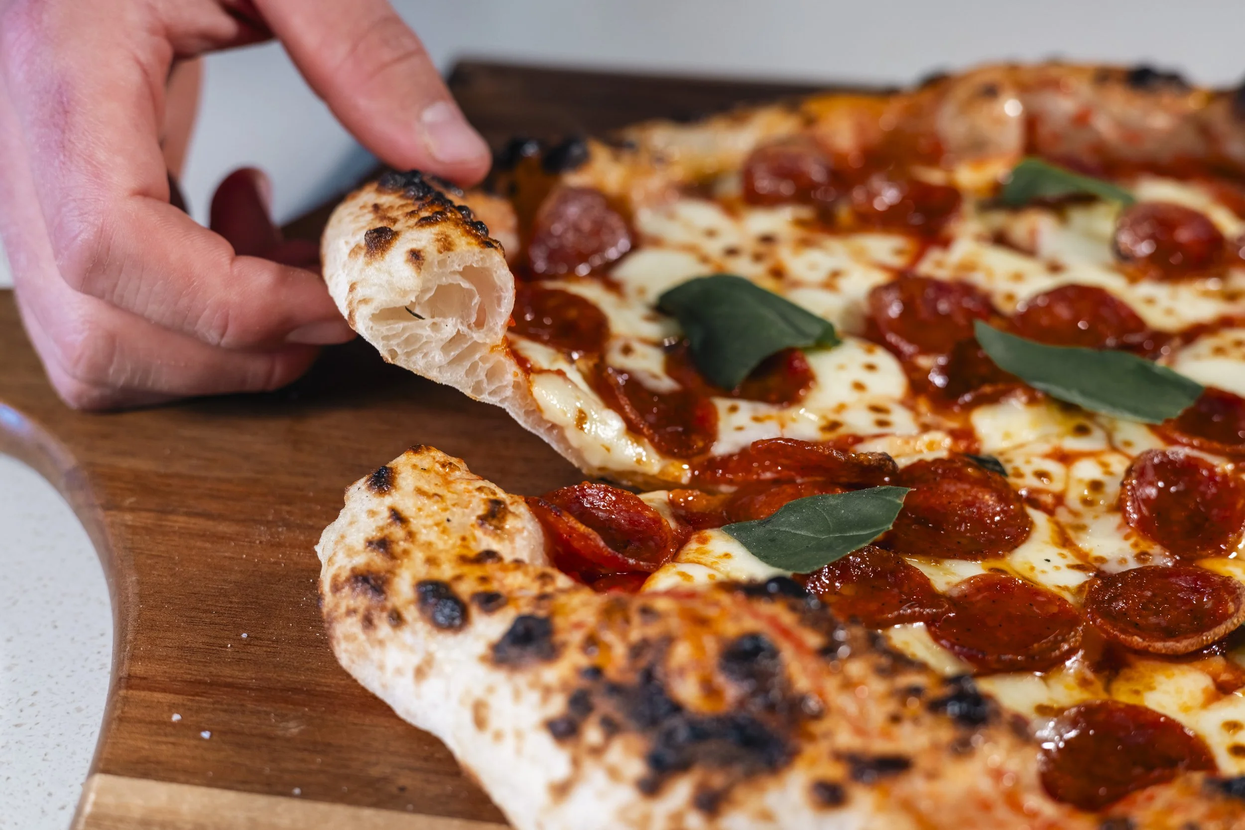 Close-up of a hand holding a piece of pepperoni pizza with melted cheese and basil on a wooden serving board.