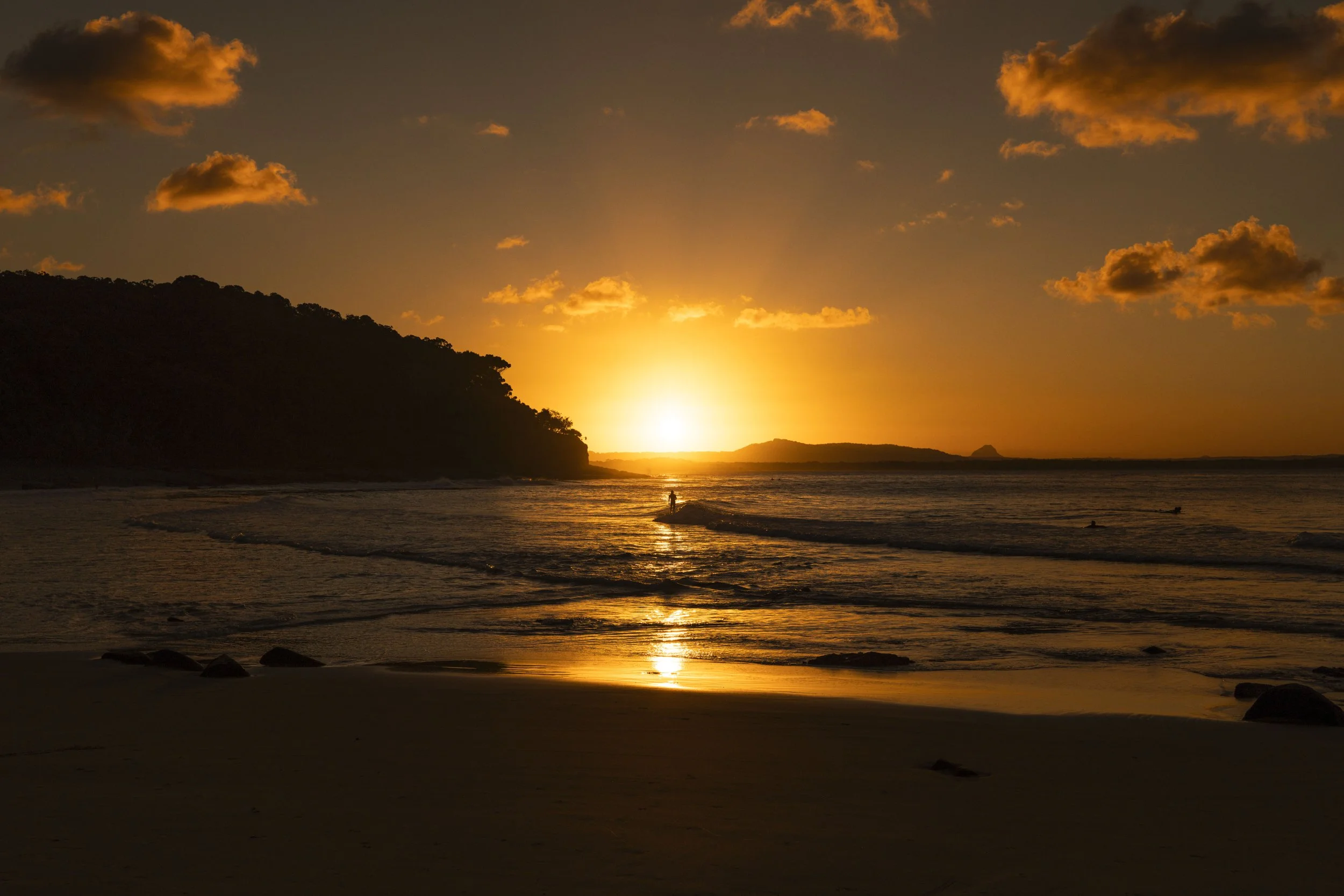 A sunset over the ocean with orange clouds in the sky, a silhouetted coastline on the left, and a person walking along the beach near the water.