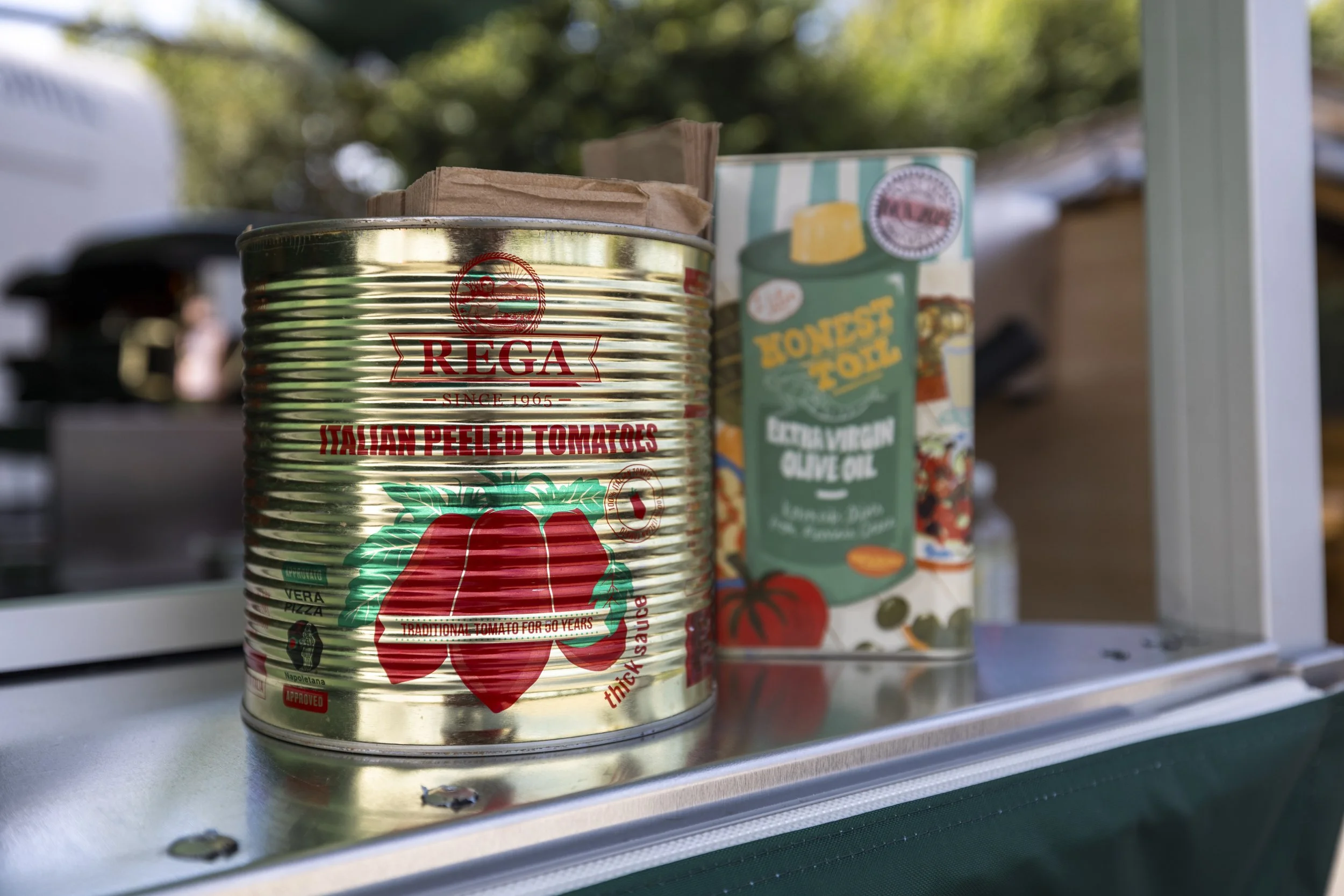 A can of REGA Italian peeled tomatoes next to a box of Honest To Goodness extra virgin olive oil on a metal shelf with a green background and sunlight.