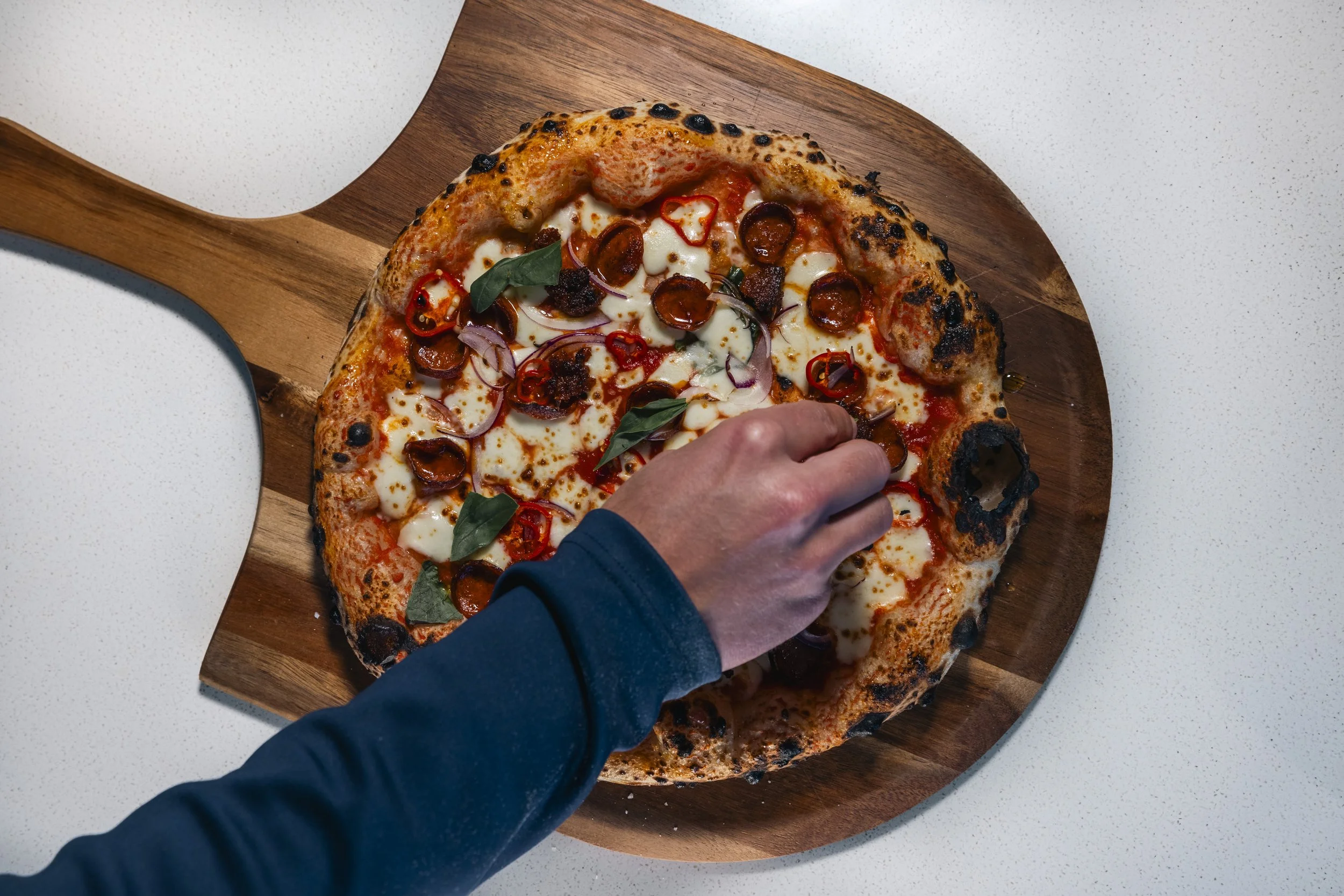 Close-up of a person selecting a slice of pizza with toppings such as pepperoni, red onions, and basil leaves, on a wooden serving board.