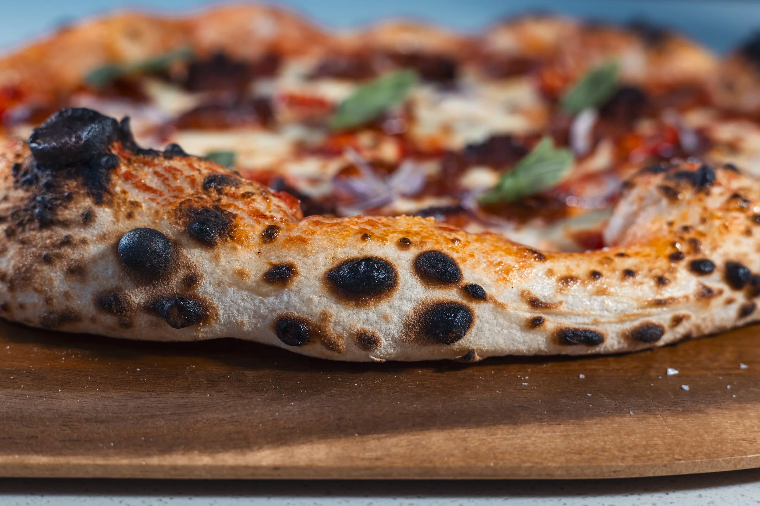 Close-up of a pizza with melted cheese, blackened spots from charring, basil leaves, and various toppings, resting on a wooden surface.