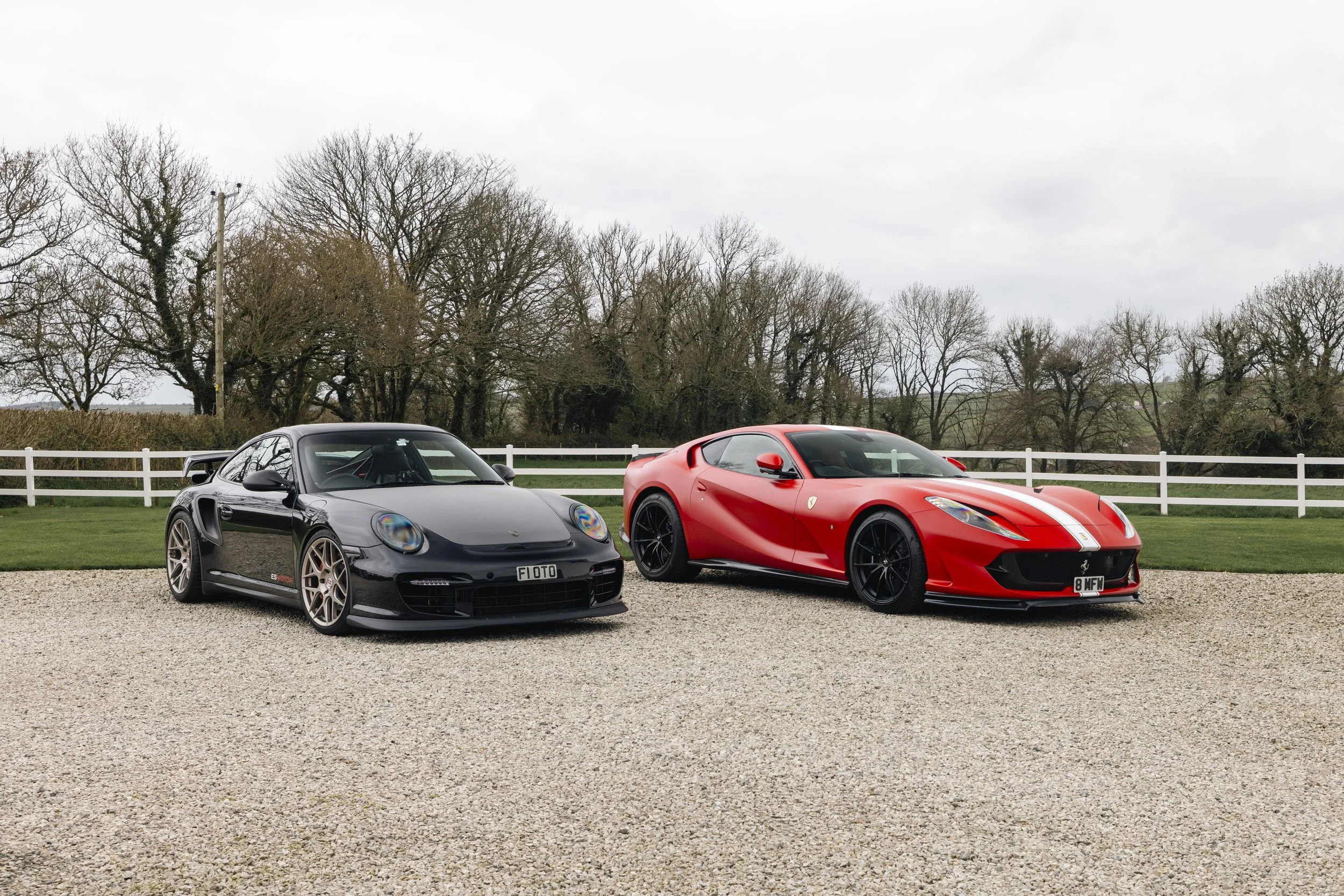 Two sports cars, a black Porsche and a red Ferrari, parked on a gravel driveway with a white fence and leafless trees in the background under a cloudy sky.