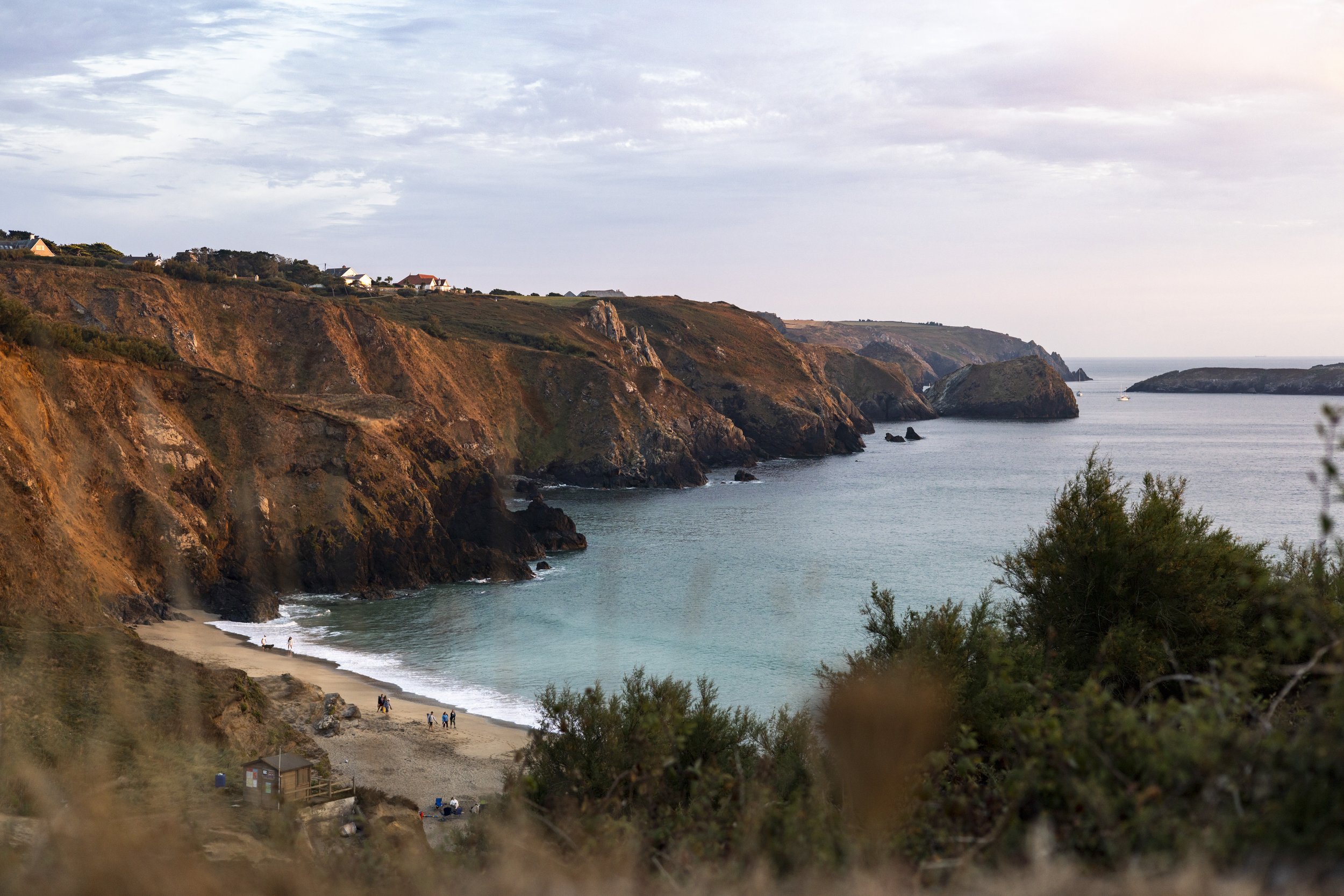 A scenic view of coastal cliffs with a small beach and people walking along the shoreline. The ocean is calm with a few boats, and houses are situated on the cliff tops under a cloudy sky.