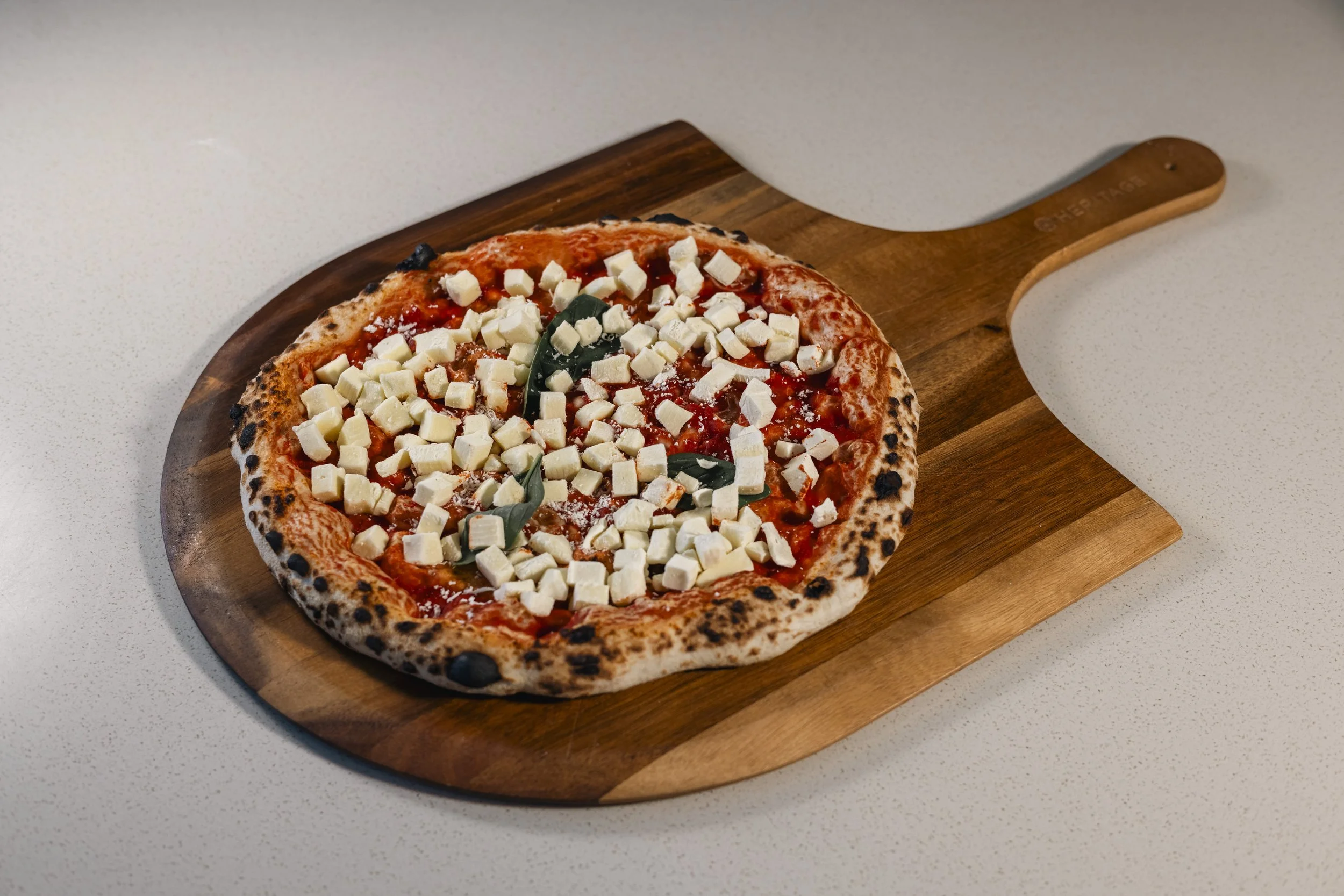 Close-up of a Margherita pizza topped with cheese and basil leaves on a wooden pizza paddle, placed on a white surface.