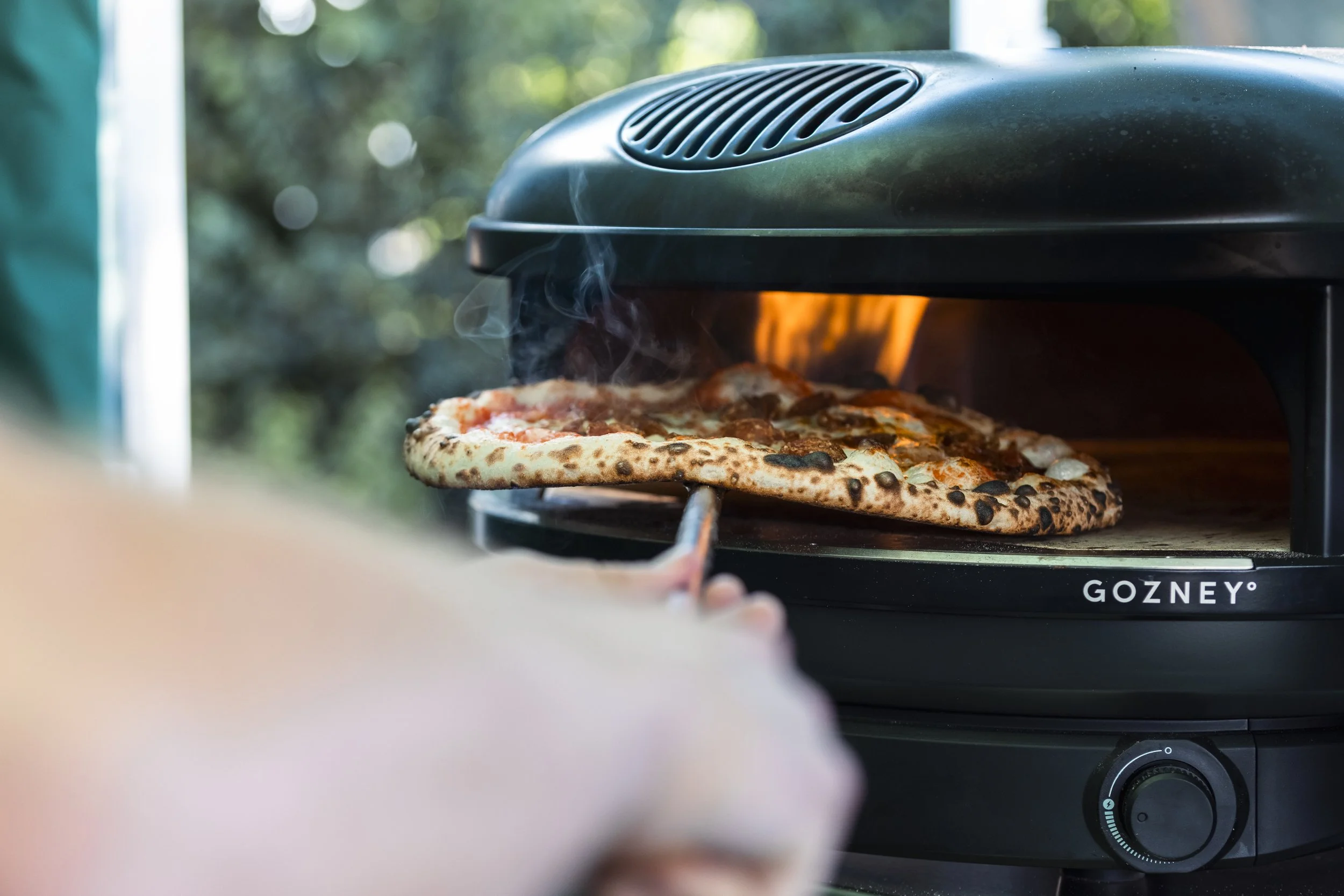A pizza baking in a black Gozney outdoor pizza oven with steam rising from it.