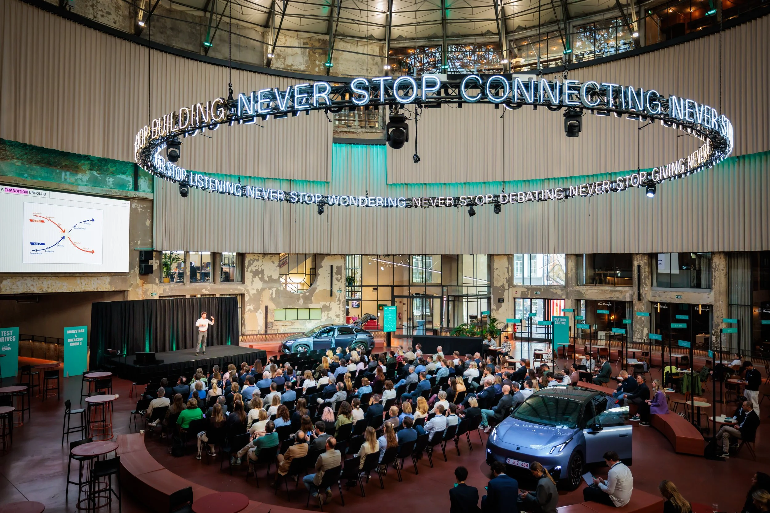 Une grande salle d'événement avec un public assis, un présentateur sur scène, deux voitures exposées, et une banderole lumineuse qui dit 'Never stop connecting, never stop building, never stop debating, never stop listening, never stop wondering, never stop giving'.