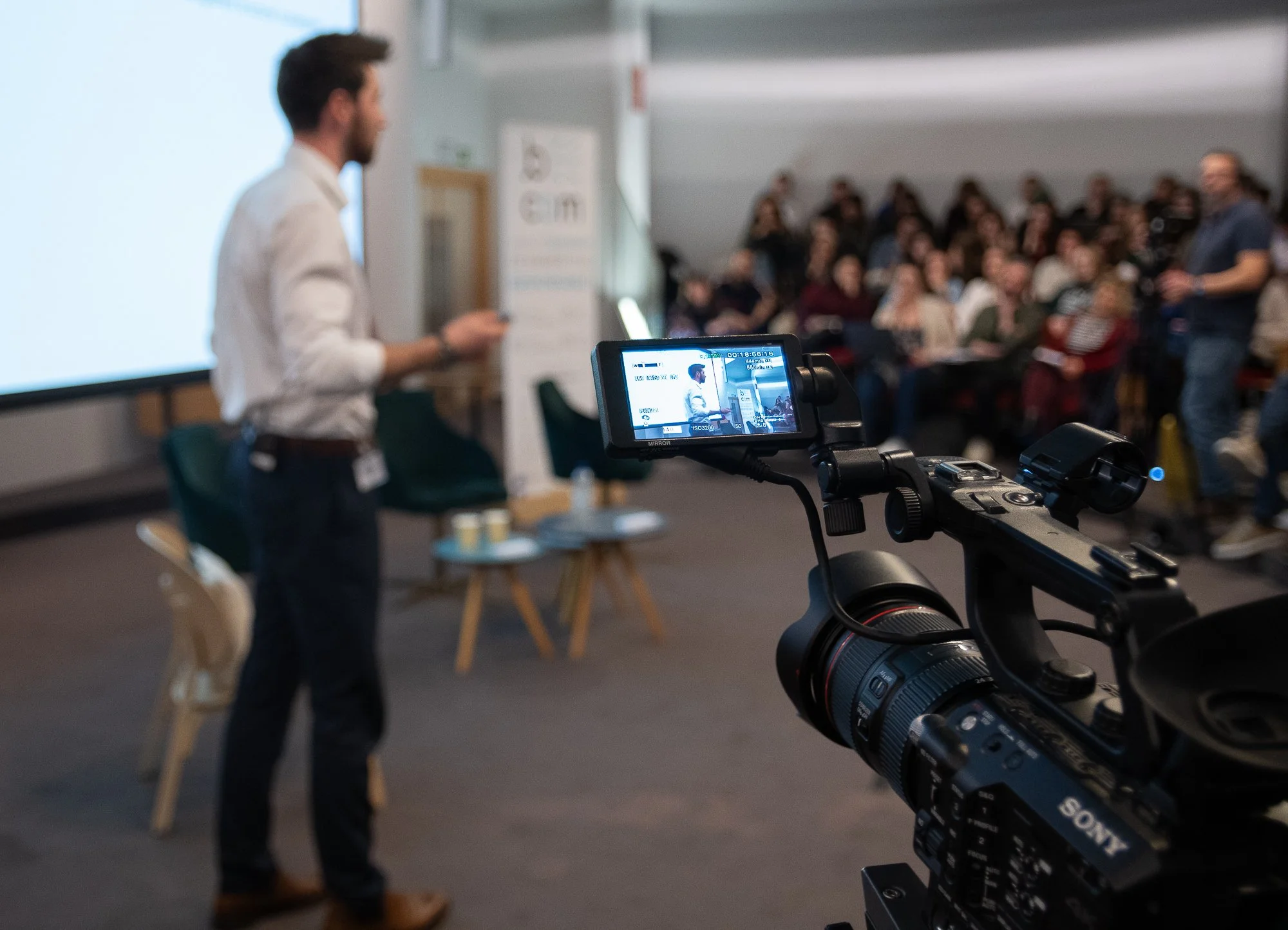 Une personne en train de donner une présentation devant un auditoire nombreux, filmée par une caméra vidéo.